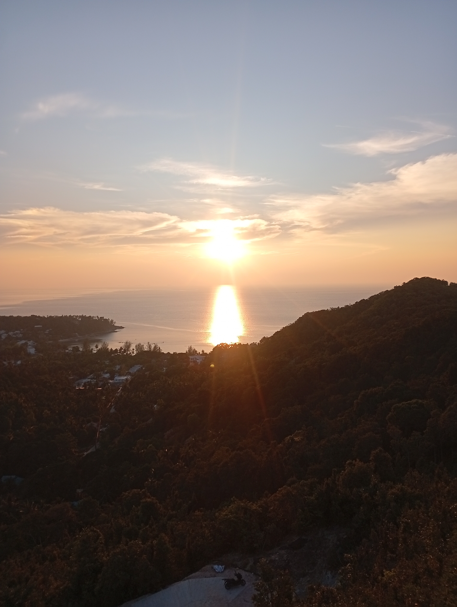 A breathtaking panoramic view captures a vibrant sunset over a coastal landscape in Baan Wang Ta Kien, Thailand. The scene is dominated by a brilliant sun positioned centrally on the horizon, casting a strong, reflective path of light across the calm water below. The sky transitions from a pale blue at the top to warm orange and yellow tones near the horizon, adorned with scattered, wispy clouds.
To the left, a curved coastline is visible, featuring a small town or resort area with white buildings and structures interspersed among lush green tropical foliage, including identifiable palm trees. The water in this bay area appears placid.
In the foreground and extending to the right, a dark, densely forested hillside or mountain slope rises, its rich green canopy appearing almost silhouetted against the bright sky and sea due to the backlighting from the setting sun. The very bottom right of the image shows a small section of a paved surface, likely a road or viewpoint, where a black scooter or motorbike is parked next to what appears to be a white sheet or blanket.
The time of day is clearly sunset, indicated by the sun's low position and the dramatic warm colors. The weather is clear and serene. No explicit human activity is captured other than the implied presence of the photographer and the parked scooter at the viewpoint. There is no visible text in the image.