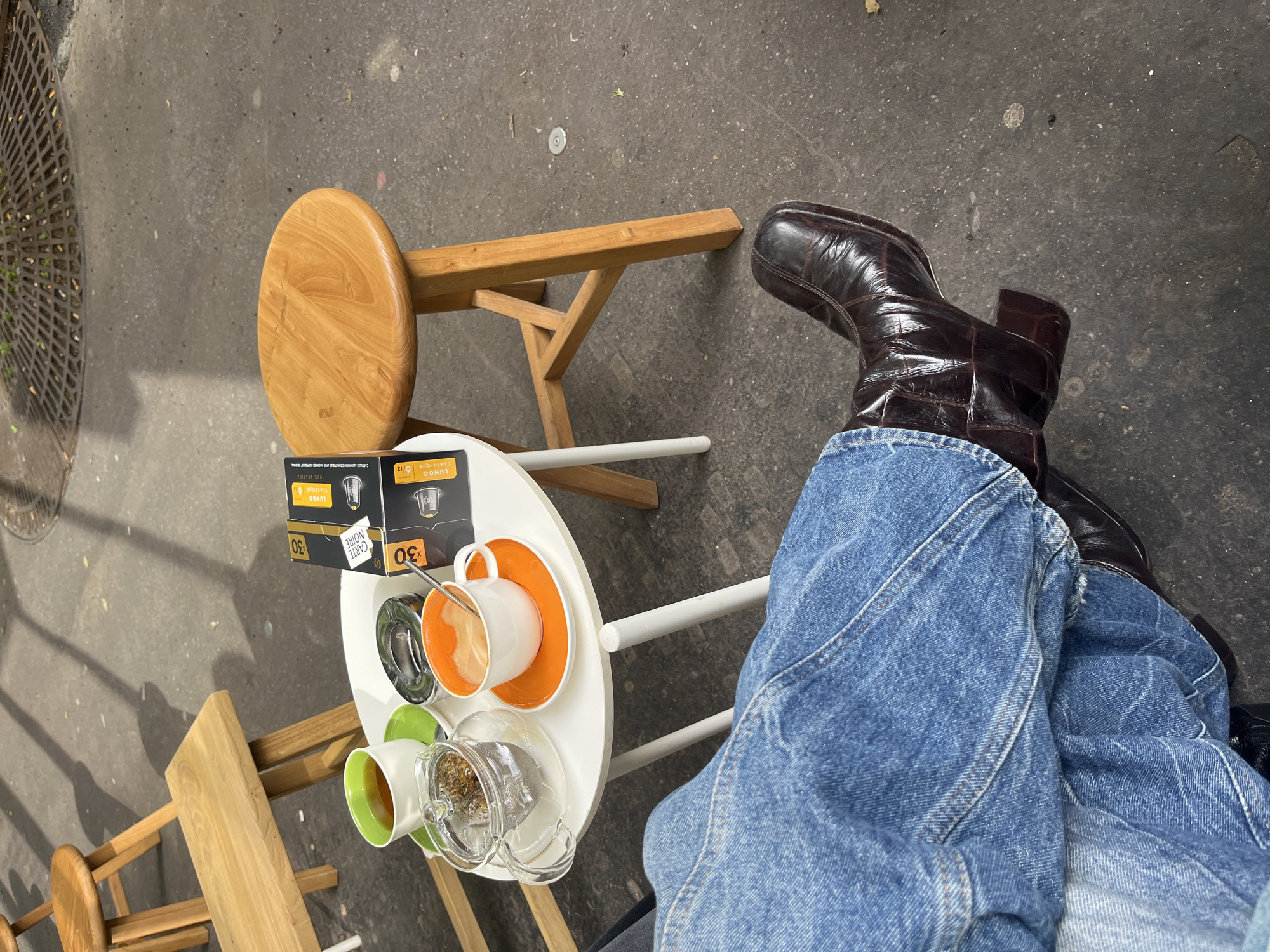 Here's a detailed description of the image:
**Overall Impression:** The image appears to be a top-down shot, capturing a casual café setting in Paris, France. The scene centers on a small white table with coffee, tea, and related items, accompanied by chairs and a person's legs and boots. The style is candid, suggesting a personal snapshot rather than a staged photograph.
**People:**
* Only the lower half of a person is visible, wearing blue jeans and dark brown leather boots. Their legs are crossed.
**Subjects/Objects:**
* **Table:** A small, round white table is the focal point.
* **Drinks:** There are at least two cups on the table - one white with an orange interior and the other white with a green interior. There's a clear glass teapot filled with what appears to be loose-leaf tea.
* **Coffee Capsules:** Two boxes of "Carte Noire Lungo Classique" coffee capsules are present, indicating the café serves this brand. The boxes mention "30 Capsules".
* **Chairs/Stools:** There are two wooden stools or chairs. One is a typical stool with a round seat. The other appears to be a shorter chair.
* **Footwear:** The person is wearing dark brown leather boots with a distinct crocodile-like texture on the leather.
* **Background Elements:** A section of street or sidewalk is visible, including a metal grate (possibly a drain cover) and the concrete surface.
**Actions/Interactions:**
* It seems likely the person is relaxing and enjoying a drink at the café.
* The placement of the items on the table suggests that coffee and tea are being consumed.
**Environment:**
* **Location:** The scene suggests a Parisian café, likely located outdoors on a sidewalk. The presence of French-language branding on the coffee capsules further supports the location.
* **Atmosphere:** The image conveys a relaxed and casual ambiance. The high-angle perspective makes it personal and intimate.
**Forensic Notes:**
* The image has been captured from an above-eye-level view, giving an overview perspective of the elements in the setting.
* The boxes of coffee capsules are placed at the edge of the table to be partially visible, indicating the camera may have been angled to capture more detail and information.
* The leather boots show some wear and tear, and the jeans have a lightly faded appearance, suggesting that the clothing is being used frequently.