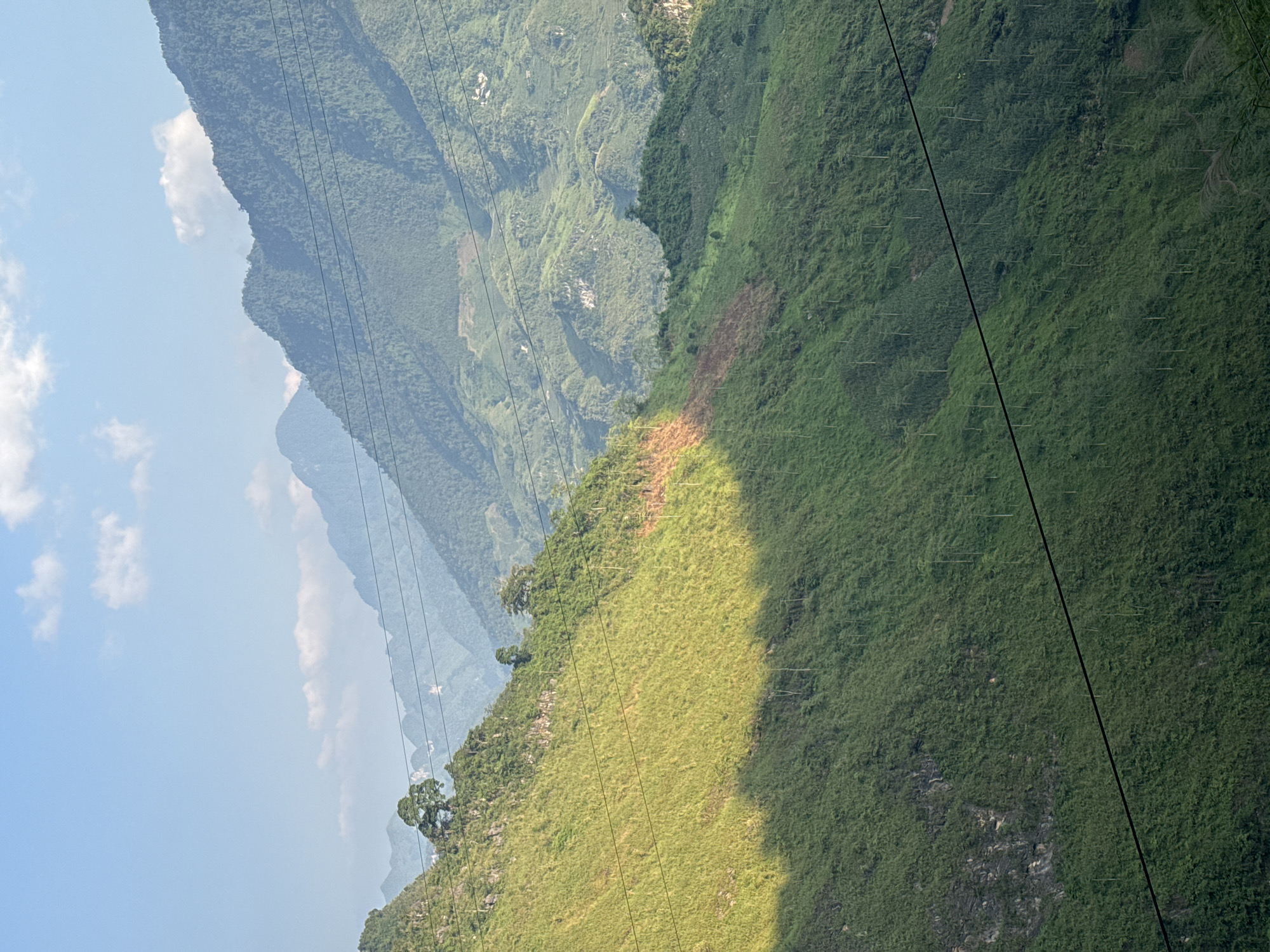 The image shows a mountainous landscape in Đường Thượng Commune, Vietnam. The scene features layers of mountains receding into the distance under a blue sky with scattered clouds. The foreground and mid-ground mountains are covered in dense green vegetation, with some areas appearing lighter in color, possibly due to variations in foliage or sunlight. Power lines stretch across the view, intersecting the landscape. The background mountains appear hazy and less defined, suggesting distance. The lighting indicates it is daytime.