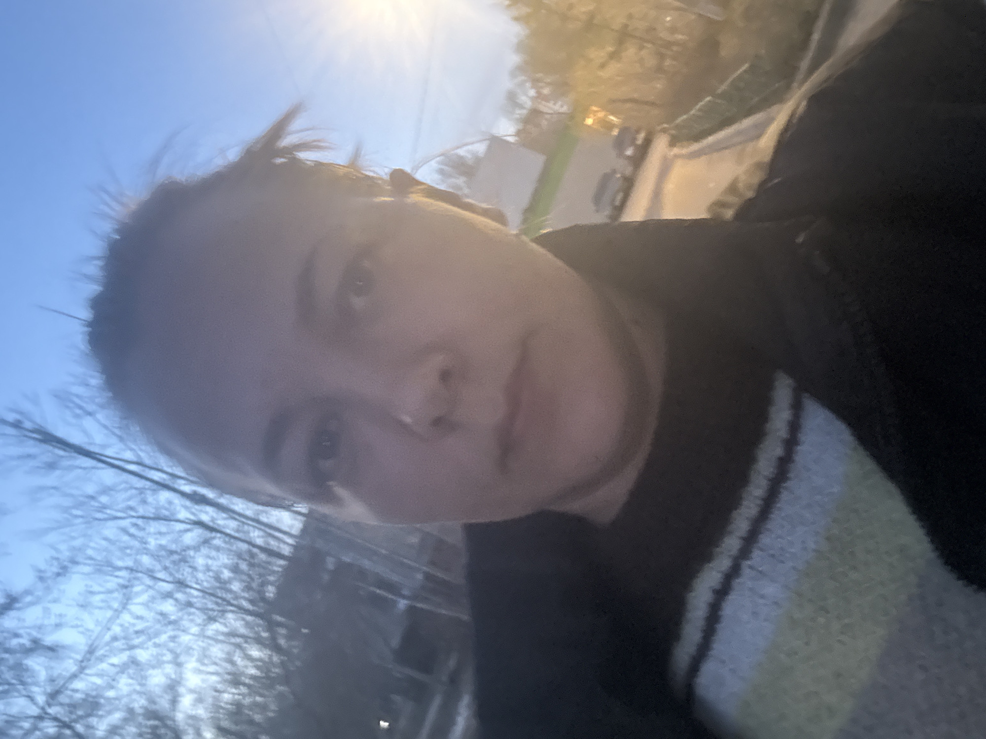 A close-up, tilted selfie taken outdoors shows a person with fair skin. The person has short hair and is wearing a black and striped sweater. The background includes a blue sky, bare tree branches, a road, and buildings. The lighting suggests it may be early morning or late afternoon. I cannot determine the location to be in Dmitrov, Russia, with the provided image.