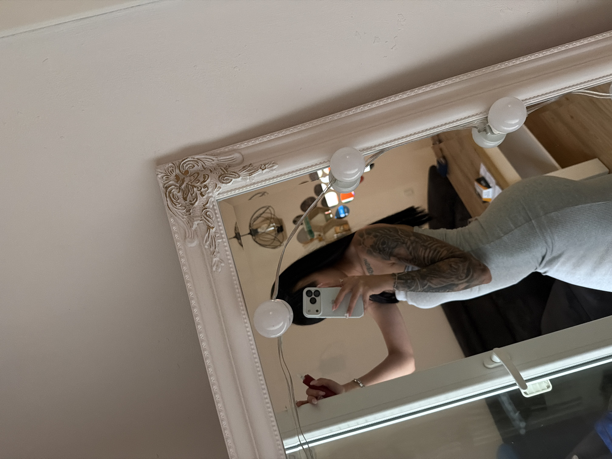 A woman with long, dark hair and tattoos on her arm is standing in front of a large, ornate mirror. She is wearing a light gray strapless dress and holding a red object in her left hand. She is taking a selfie with a white smartphone. The mirror is surrounded by a string of round lights. In the reflection of the mirror, a wooden table with some items on it and a dark sofa can be seen. The scene appears to be an indoor setting, possibly a bedroom or dressing room, with a pale wall in the background. The location is not specified as Plovdiv, Bulgaria.