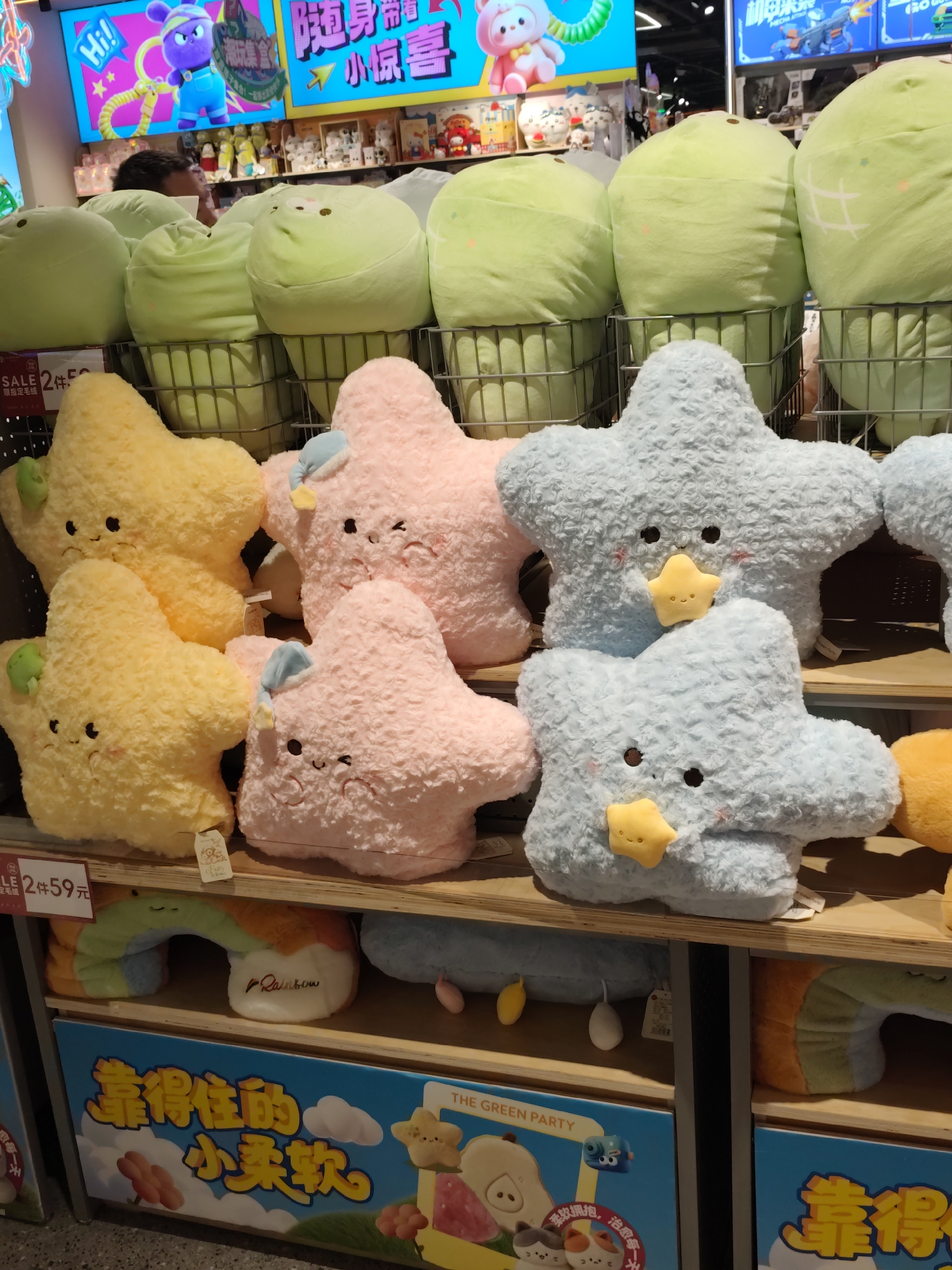 The image shows a retail display of plush toys in Daqing, China. The toys are arranged on shelves and include several star-shaped pillows in yellow, pink, and light blue, as well as large, round green plush toys. Some of the star pillows have embroidered faces and are wearing small hats. A large sign with Chinese characters is visible at the bottom of the image, along with other decorative elements like clouds and fruit. The background includes more merchandise and bright lighting, suggesting a store environment. A person's head and shoulder are partially visible in the upper left portion of the image, looking towards the display.