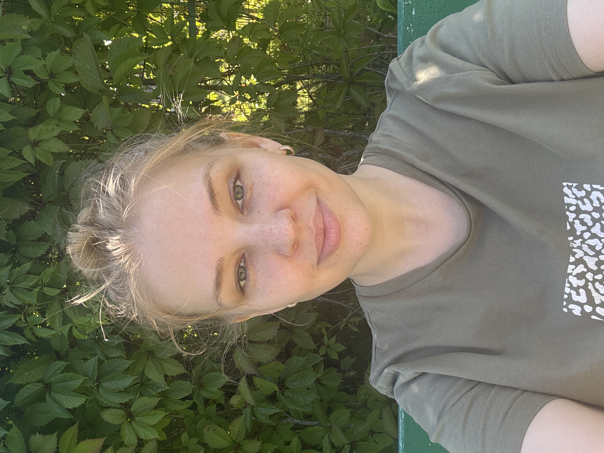 A young woman with blonde hair pulled into a bun is looking at the camera with a slight smile. She has light skin with freckles and green eyes. She is wearing a khaki t-shirt. The background consists of lush green foliage and a hint of a green bench. The image appears to be taken outdoors during the daytime, possibly in the city of Dunilovo, Russia, given the presence of dense vegetation.
