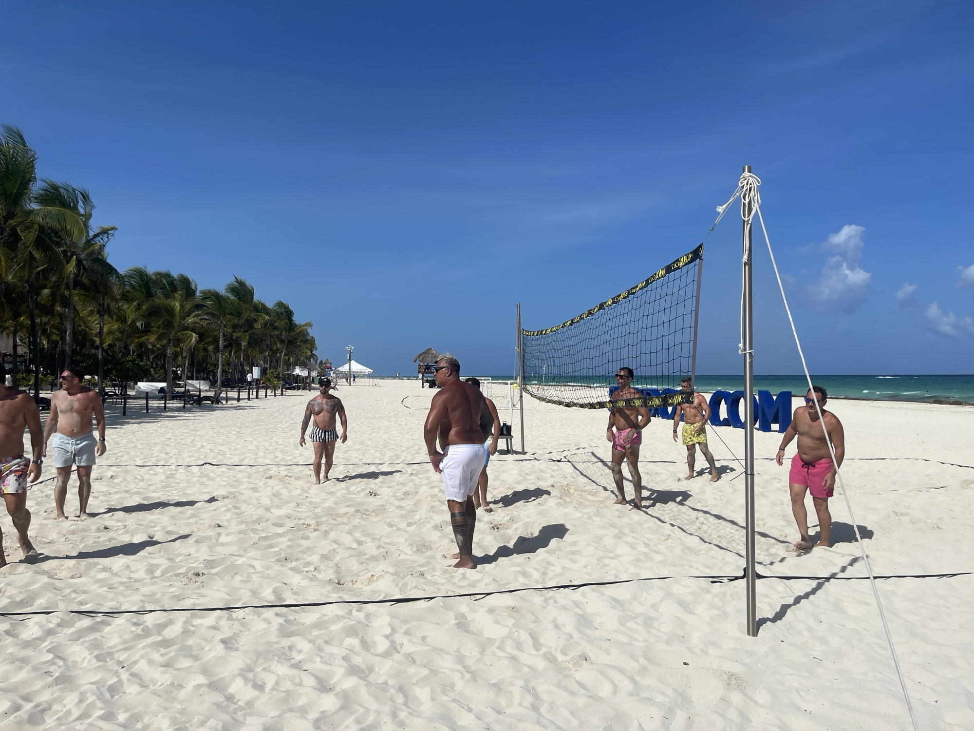 Several men are playing beach volleyball on a sunny day at a beach in Playa del Carmen, Mexico. The beach has white sand and is lined with palm trees. A volleyball net is set up on the beach. The men are wearing swim trunks and are of varying builds and ages. Some are standing and ready to play, while others are actively engaged in the game. Behind the volleyball net, partially obscured by the net and the players, are a row of large blue letters spelling "OCCIDENTALCOMS". Beyond the beach are the ocean and a clear blue sky. In the background, there are some beach chairs and what looks like a thatched-roof structure.