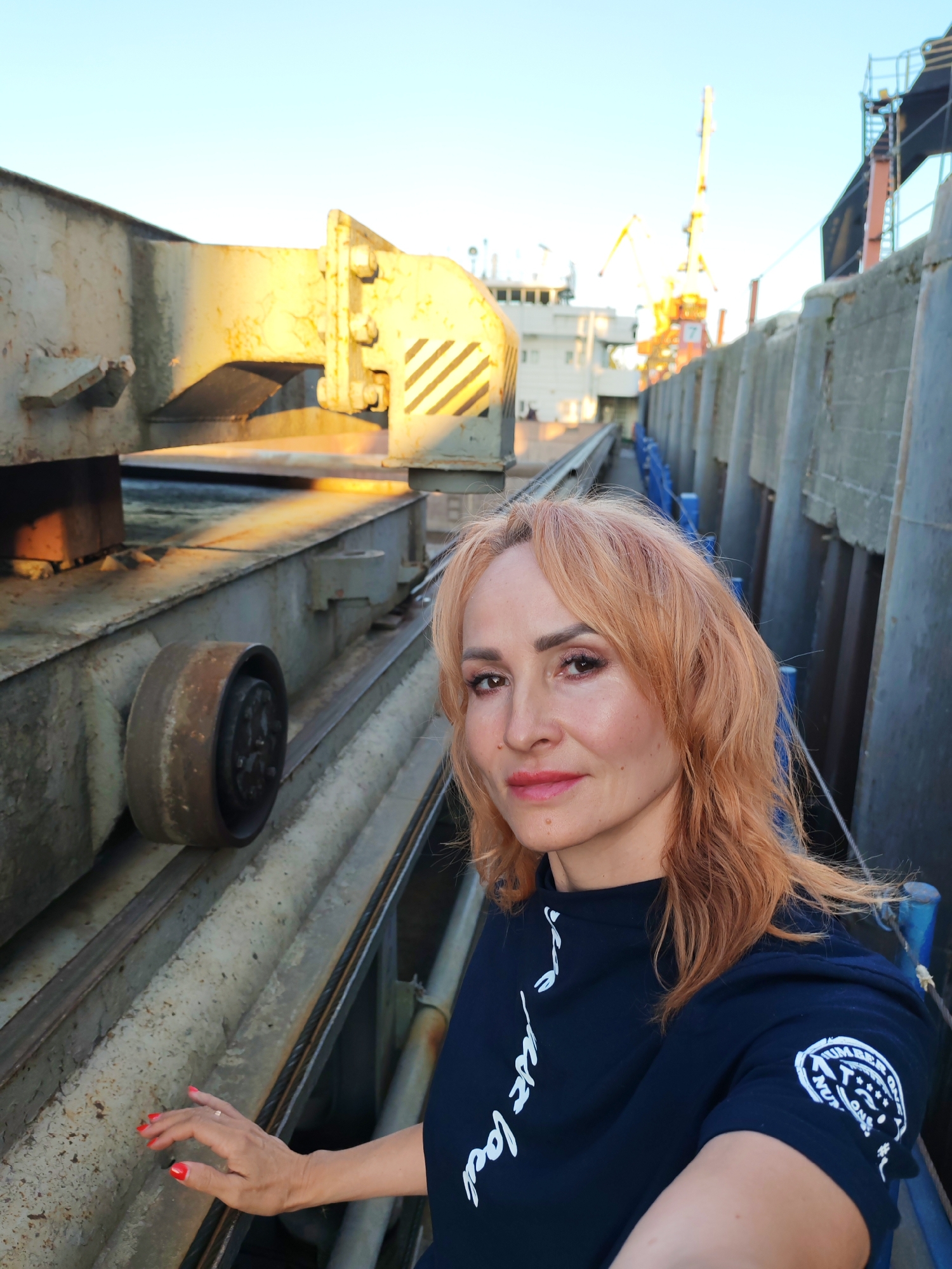 A woman with auburn hair is taking a selfie on a sunny day, with a clear blue sky overhead. She is wearing a dark blue shirt with white lettering and a circular emblem on the sleeve. Her left hand, with bright red nail polish, rests on a metal railing. The background shows a pier or dock area with industrial elements, including large metal structures, a metal wheel on a track, and distant cranes and a ship. The setting appears to be a port or industrial zone, potentially in Rybinsk, Russia, given the context. The overall impression is one of a casual moment captured in an industrial waterfront environment.