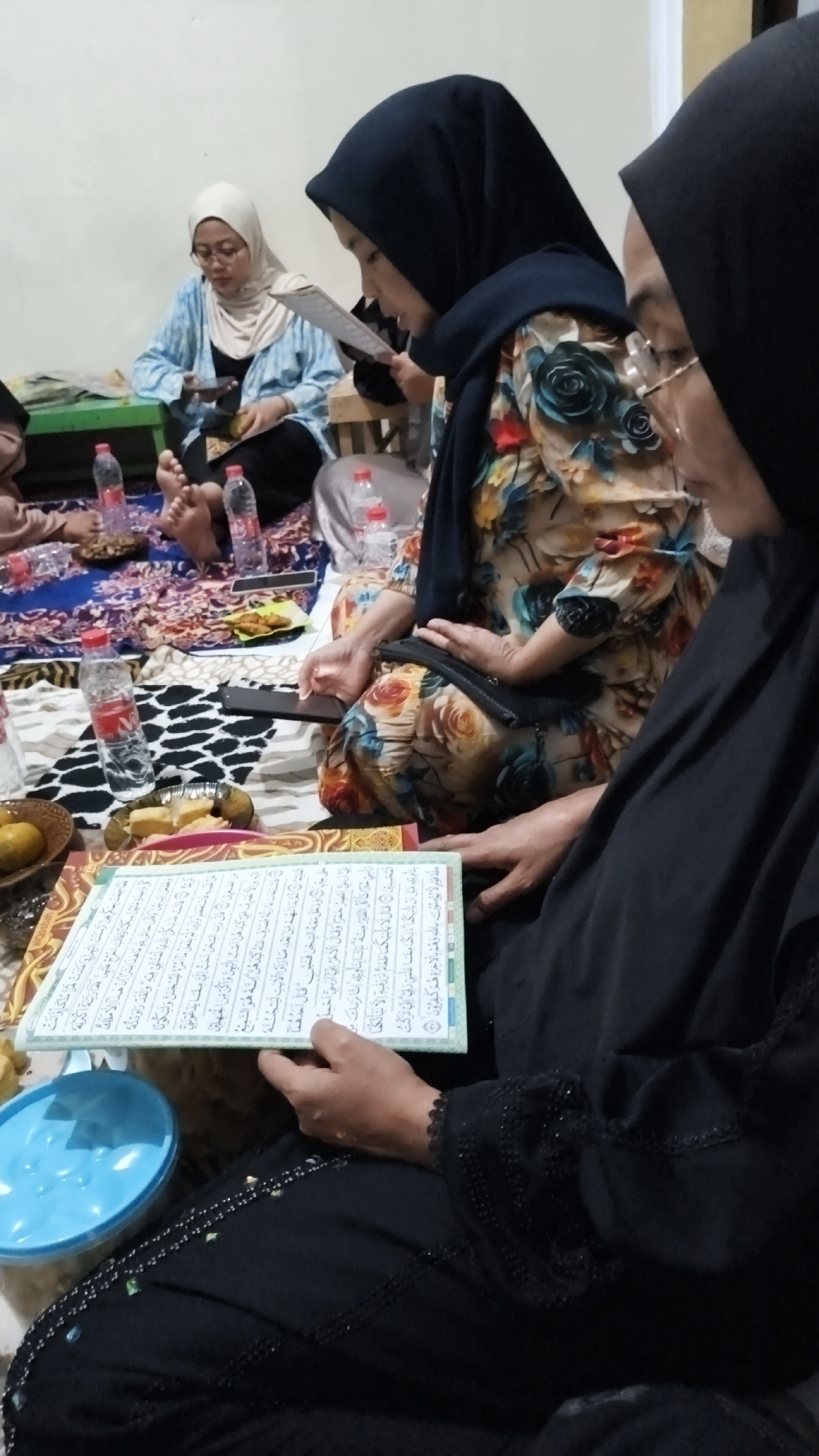 In Dalegan, Indonesia, three women are seated on the floor, engaged in reading from a Quran. Two women in the foreground are wearing black hijabs, with one of them also wearing a black abaya. The third woman in the background is wearing a light-colored hijab and a blue and white patterned top. Scattered around them are several bottles of water, a plate of snacks, and a blue bowl. The scene appears to be indoors, possibly a home, with a plain wall in the background and a patterned rug covering the floor. One of the women in the foreground is holding a mobile phone.