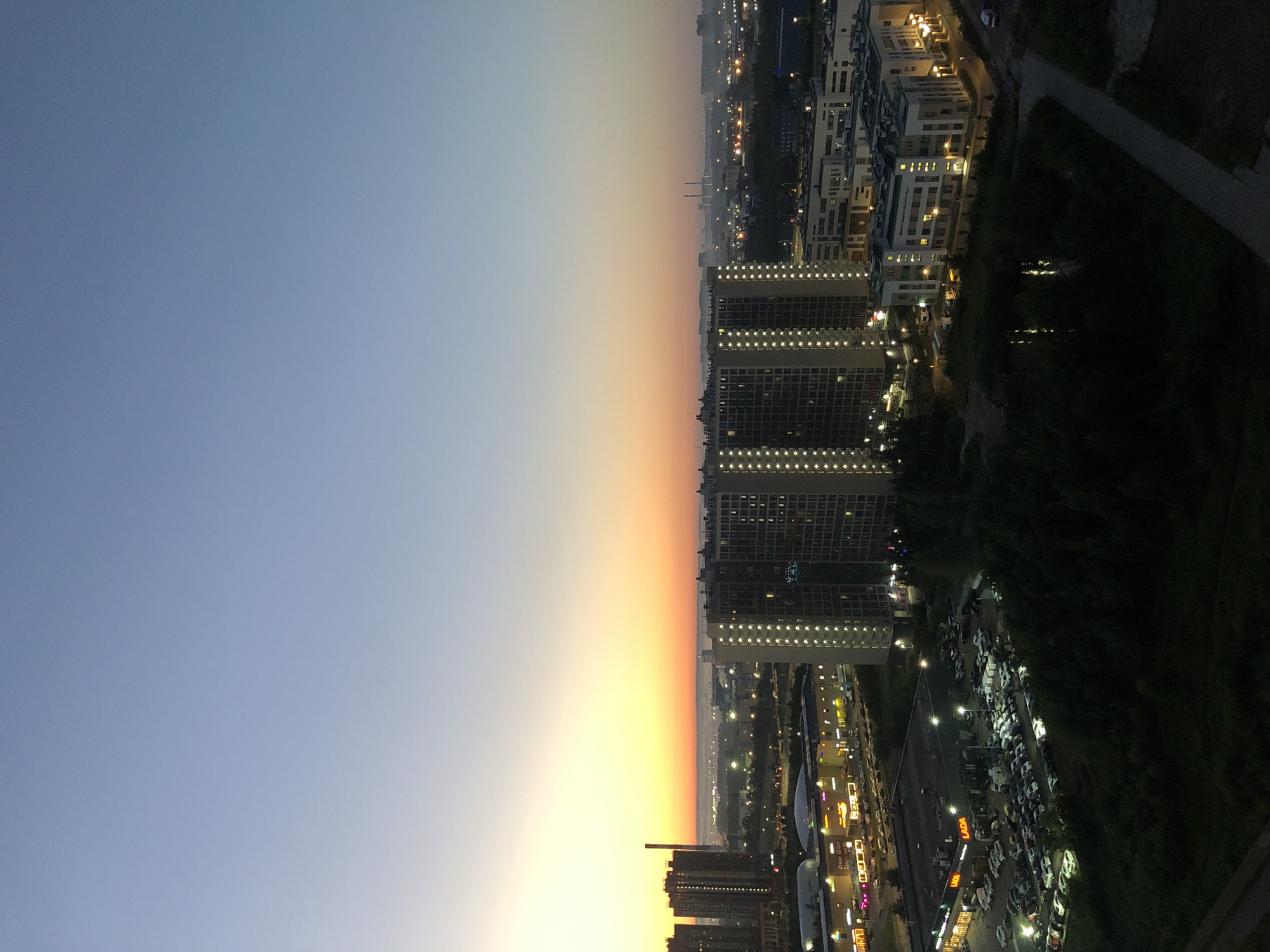 An elevated, wide-angle, vertical-format, full shot captures a cityscape at dusk. The sky transitions from a pale blue at the top to a soft orange and yellow gradient near the horizon, indicating either sunrise or sunset. In the foreground, silhouetted trees and a small pond are visible. To the left, a large, modern apartment building with numerous illuminated windows stands prominently. Adjacent to it, a shopping center with brightly lit storefronts and a large parking lot filled with cars are apparent. In the mid-ground, another apartment complex, also illuminated, is situated. The background reveals a hazy industrial or urban landscape with distant buildings and what appear to be smokestacks or industrial towers. The overall impression is of a developing urban area in the twilight hours.
