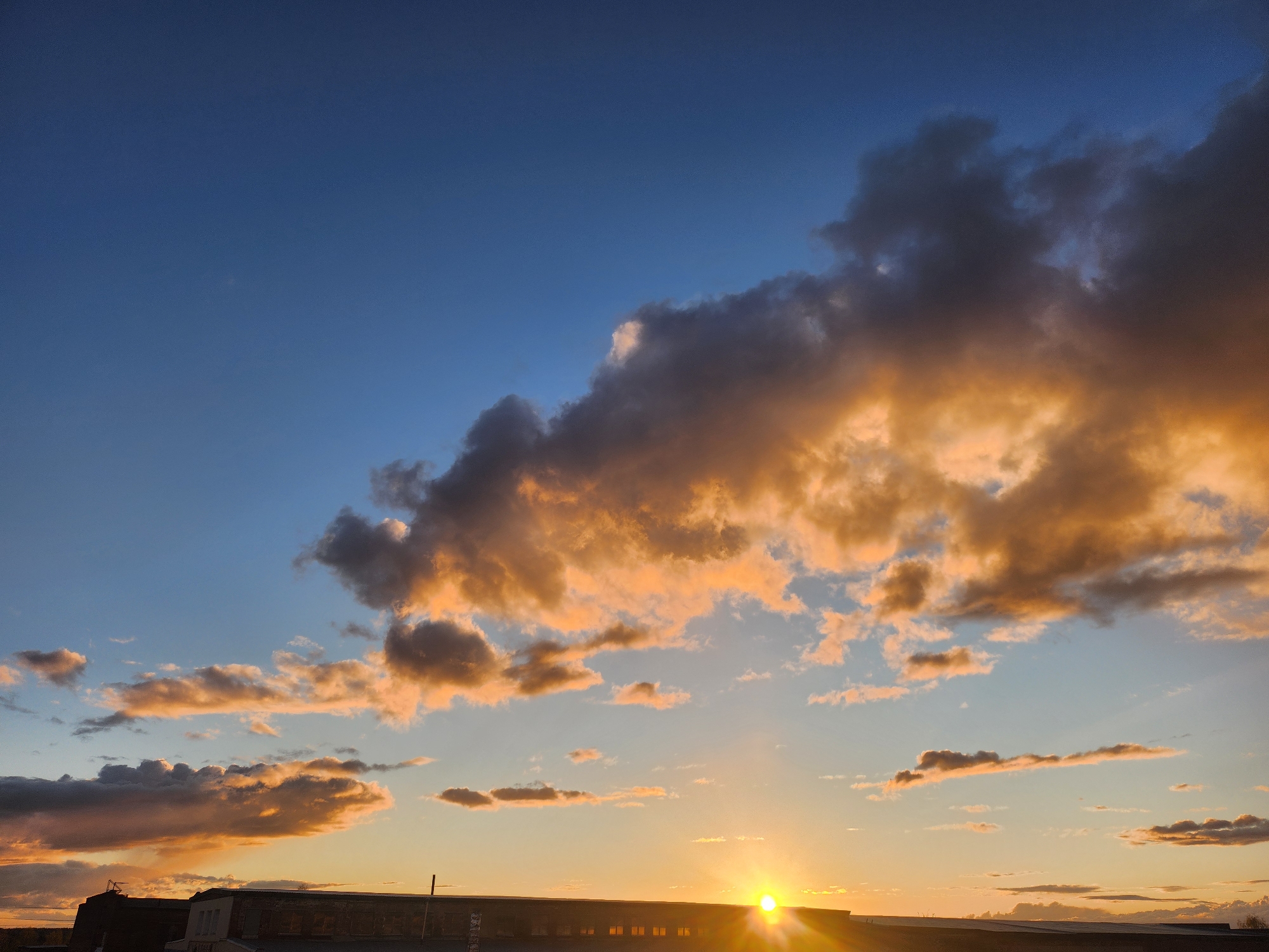 The image shows a sunset over a building in Krasnogorskoye, Russia. The sky is a gradient of blue to orange, with clouds illuminated by the setting sun. The building is dark and silhouetted against the bright sky.