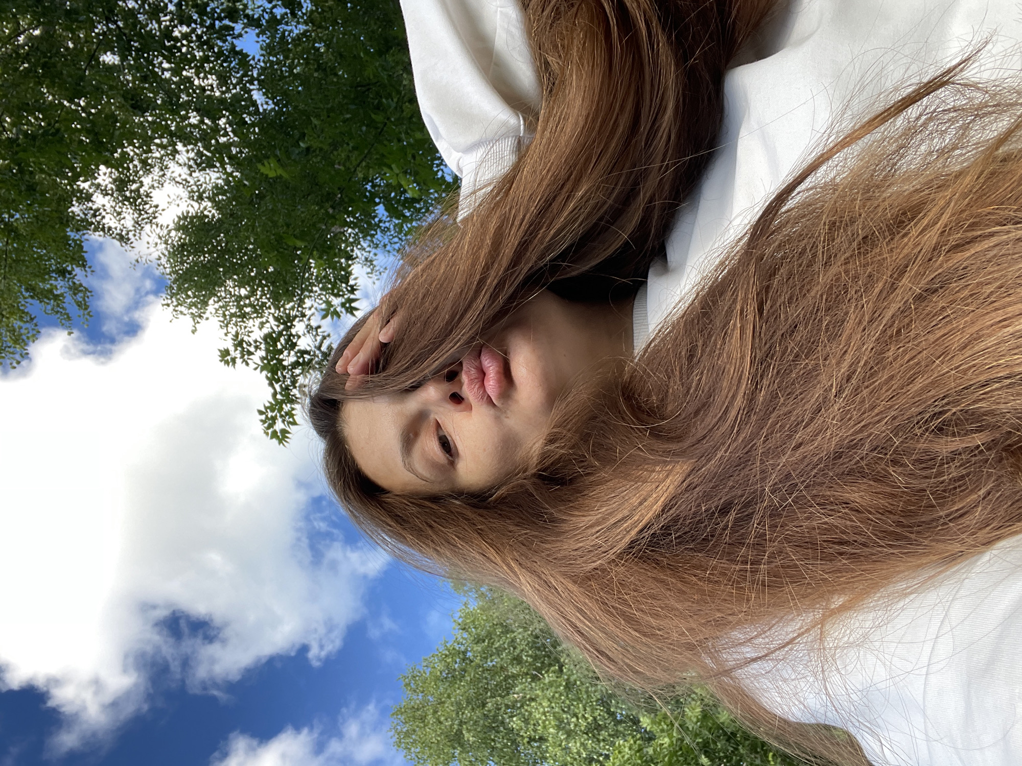 The media file is a selfie taken outdoors with a bright, cloudy sky and green foliage in the background. The subject is a young person with long, brown hair, wearing a white hoodie. They are lying down, with their head angled towards the camera, and their hair is spread out around them. Their lips are pursed in a "duck face" pose, suggesting a playful or whimsical mood. The lighting indicates it is daytime, likely sunny with scattered clouds. The foliage and blue sky suggest a natural outdoor setting, possibly a park or garden. There are no visible text elements in the image.