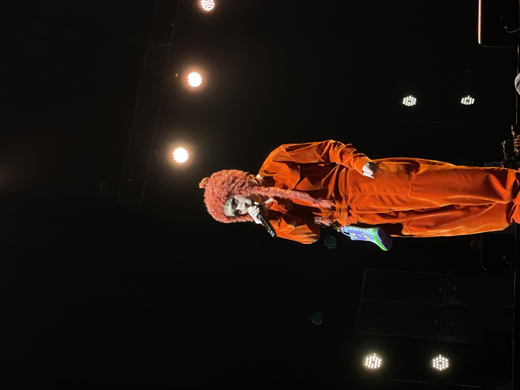 A person wearing an orange jumpsuit with a furry orange hood is holding a microphone and performing on a stage. The setting is dark, with multiple spotlights illuminating the performer. Speakers are stacked behind the person. The exact location in Salembaran, Indonesia, cannot be determined from the image.