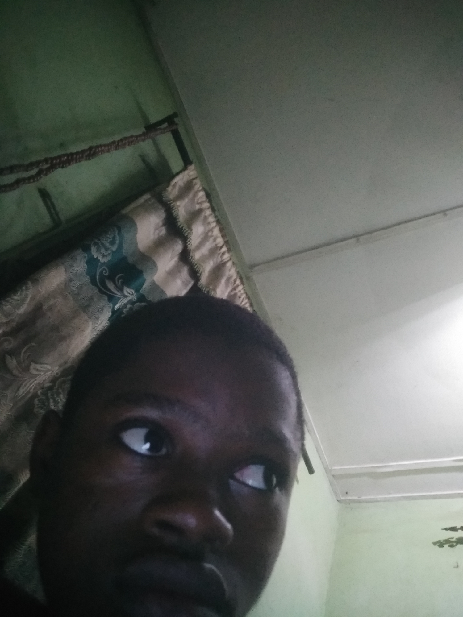 The image is a close-up selfie of a dark-skinned person with short hair, looking to the side with one eye visible. The background consists of a light green wall and a white ceiling. A curtain with a floral pattern is partially visible on the left. The location is unknown; there is no visual evidence to suggest it is Shangisha, Nigeria.