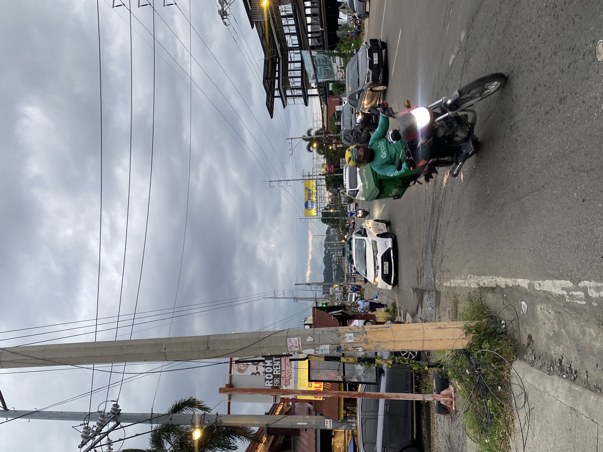 The image depicts a street scene in Davao City, Philippines, likely during overcast weather given the cloudy sky. In the foreground, a man wearing a green long-sleeved shirt with "Grab" logo and a helmet rides a motorcycle with a green insulated delivery box on the back. The motorcycle's headlight is illuminated. The man is in motion, with the motorcycle's front wheel slightly angled.
The street is paved with asphalt and has lane markings. To the left of the motorcycle, a gray car is parked on the side of the road, and beyond it, a building with a red roof and a sign that reads "ROOM FOR RENT" is visible. Several utility poles with numerous wires are prominent throughout the scene.
Further down the road, several other vehicles are present, including a white sedan with a license plate that is partially visible, a dark-colored SUV, and other cars. Pedestrians are also seen walking on the sidewalk. The background shows buildings, more utility poles, and distant hills. The overall impression is a busy urban environment with ongoing commercial and transportation activities.