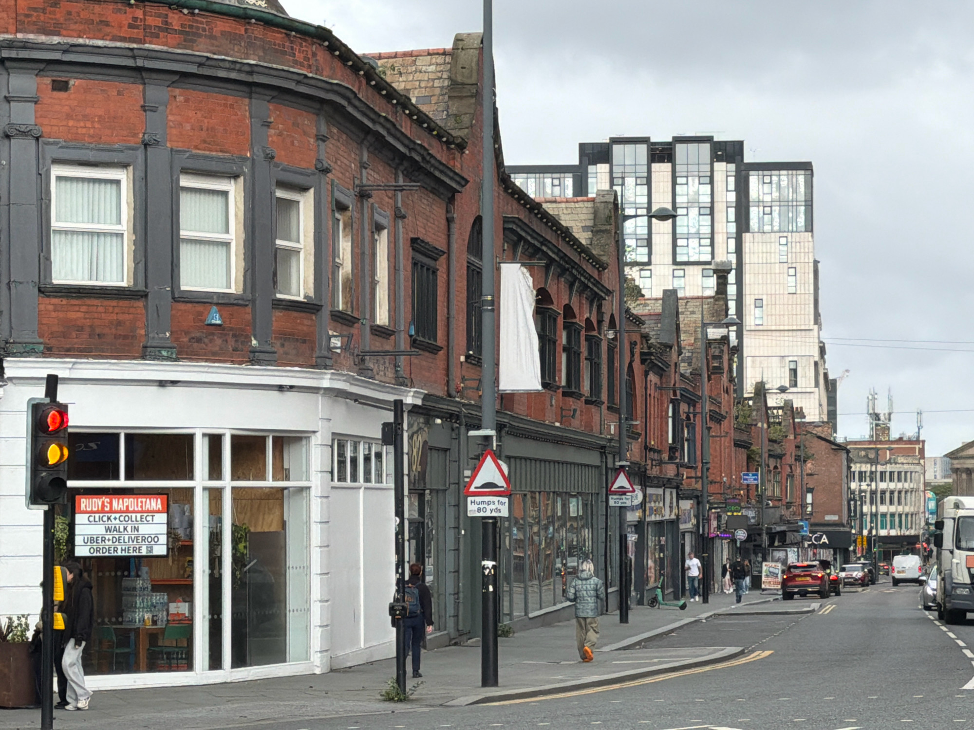 The image depicts a street scene in a city. In the foreground, a red brick building with grey detailing has multiple windows and a white storefront. A traffic light with red, yellow, and green signals stands to the left of the building, next to a planter. A sign on the storefront reads "RUDY'S NAPOLETANA CLICK+COLLECT WALK IN UBER+DELIVEROO ORDER HERE". Two individuals are standing on the sidewalk in front of the building.
The street continues into the background, lined with more buildings of varying architectural styles and heights. A modern, multi-story building with a glass facade is visible further down the street, contrasting with the older brick structures. Traffic signs, including a speed bump warning sign, are posted along the sidewalk. Several cars are driving on the road, and a white van is parked on the right side of the street. People are walking on the sidewalk. The sky is overcast.