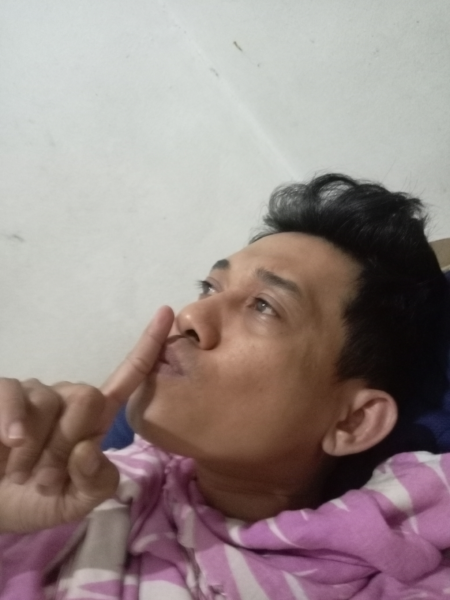 A man is resting, possibly in a bed, with a pale wall as the background. He is wearing a pink and white striped garment, possibly a shirt or blanket. His right hand is raised to his lips, with his index finger gently touching his mouth, suggesting a gesture of silence or contemplation. He is looking upwards and to the right, his eyes slightly open. The lighting appears to be from an artificial source. The scene is a close-up shot, focusing on the man's face and upper body. There is no identifiable location in Batam, Indonesia, as the environment is generic.