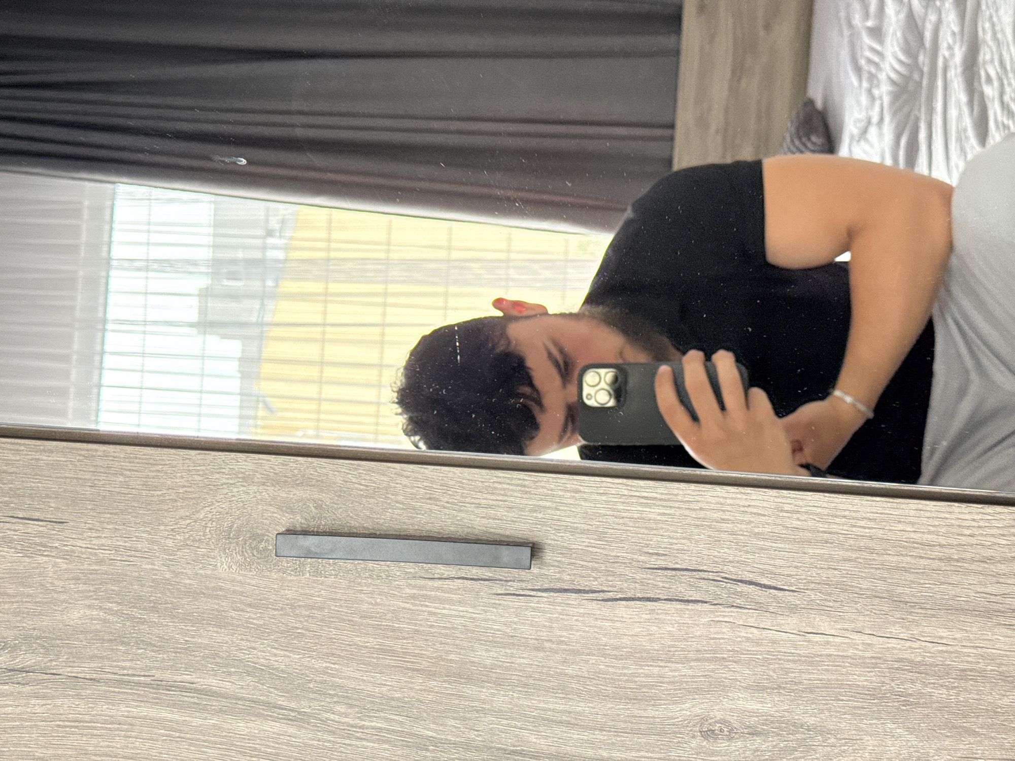 The media file captures a reflection of a person taking a selfie in what appears to be a mirror or reflective surface, positioned above a wooden furniture piece.
**Subjects:** The main subject is a person, reflected in the surface. They have dark, curly hair and are wearing a black short-sleeved t-shirt. A glimpse of their ear shows a reddish spot, possibly an earring. They are holding a dark-colored smartphone, with multiple camera lenses visible, to take the photo. A white bracelet is visible on their left wrist. Their upper arm shows notable muscle definition. The lower half of their body, reflected, suggests they are wearing light-colored, possibly grey or white, bottom wear.
**Objects:** The reflective surface itself shows small specks of dust and a distinctive small, white, comma-shaped mark near the top center. Below the reflection is a wooden surface, likely a dresser or table, characterized by a prominent light-brown grain pattern. It features a sleek, horizontal black handle. In the background of the reflection, seen through what appears to be a window or opening, are architectural structures with horizontal elements, possibly blinds or building facades, under a bright sky.
**Activity:** The person is actively engaged in taking a mirror selfie, holding their phone in a way that captures their reflection.
**Notable details:** The scene is brightly lit, suggesting it is daytime. The reflection reveals an outdoor environment, though specific location cues for Varna, Bulgaria, are not present. The furniture's texture and modern handle are clearly visible. There is no visible text in the image. The overall impression is that of an indoor setting with a view to the outside, captured through a mirror.