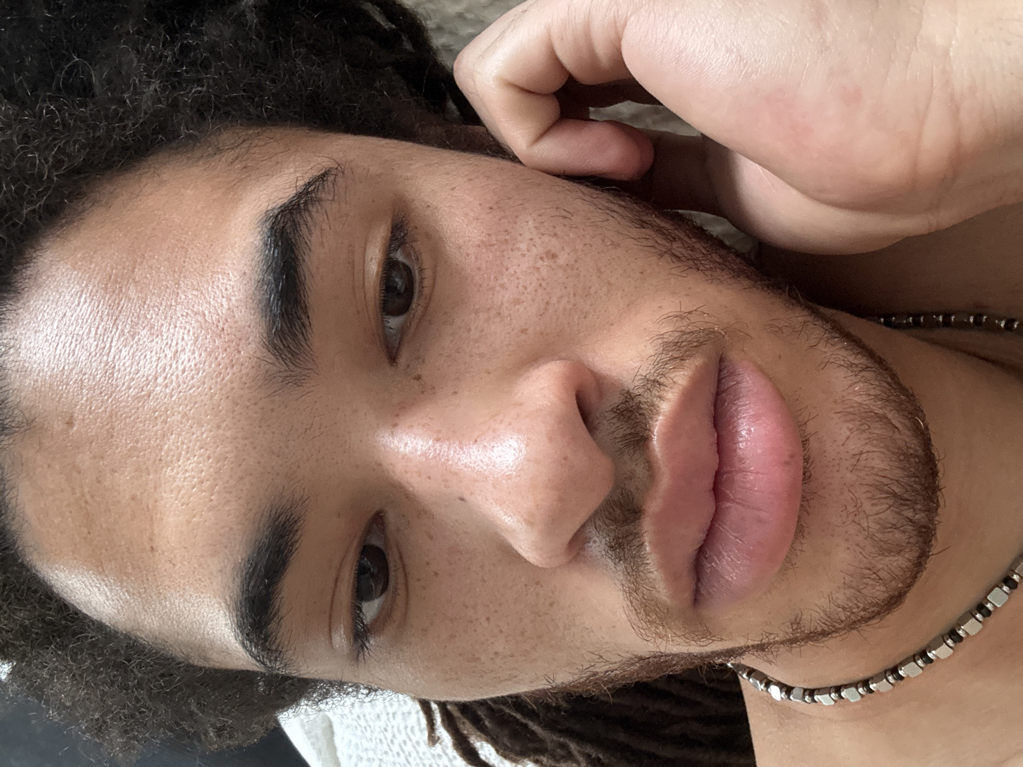 A close-up, portrait-style photograph features a person with dark curly hair and brown eyes. The individual has a visible beard and freckles on their face. Their right hand is positioned near their right ear. They are wearing a beaded necklace, and the background is slightly blurred and indistinct. No evidence indicates a location in the city of Paris, France.