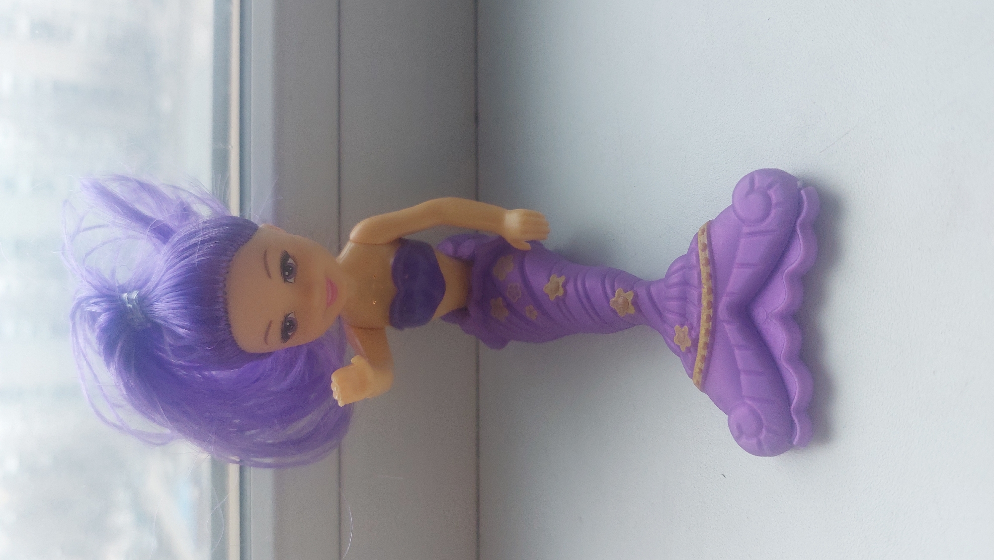 A purple plastic mermaid doll lies horizontally on a white surface near a window. The doll has purple hair, a purple top, and a purple tail with gold floral accents. The window reveals an out-of-focus cityscape, suggesting an interior location. It is not possible to determine if the location is in the city of Novosibirsk, Russia.