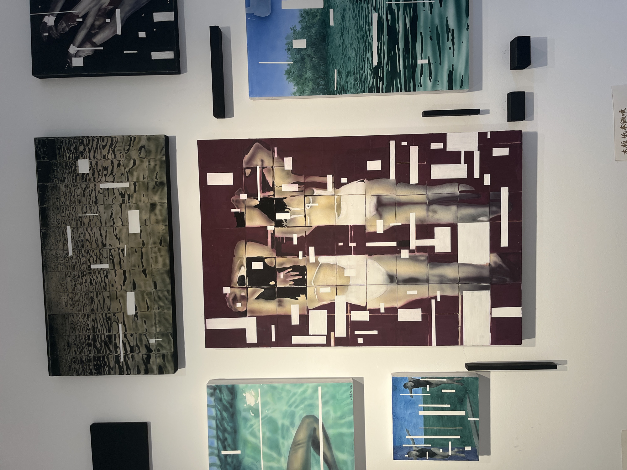 The media file displays a collection of artwork mounted on a white wall, likely in a gallery or exhibition space. The artworks are varied in style and subject matter. In the upper left, a framed piece shows a close-up of legs, possibly human, with red accents, rendered in black and white with white rectangular elements interspersed. To its right is a large, horizontally oriented painting depicting a landscape with trees against a blue sky, transitioning into a green, abstract water-like pattern with white vertical and horizontal lines.
Below these, a large rectangular painting with a dark burgundy background features two figures, appearing to be women, in what looks like a poolside setting. They are depicted in a fragmented or deconstructed style, with white geometric shapes overlaying their forms. To the left of this piece is another large, vertical artwork with a dark, textured background, also adorned with white rectangular and linear elements.
Below the burgundy-toned painting, two smaller square artworks are visible. The one on the left shows a person's legs submerged in bright green water, with white lines suggesting lanes or markings. The artwork on the right depicts swimmers in a blue and green setting, again with white lines and fragmented figures.
Several black, rectangular objects are mounted on the wall, appearing as sculptural elements or part of the mounting system for the art. A piece of paper with Chinese text is visible on the far right edge of the frame. The overall impression is one of a modern art exhibition with a focus on figurative and abstract elements, often incorporating geometric patterns. The lighting appears to be artificial, typical of an indoor exhibition. There are no clear indicators of time of day or weather. The context provided states the location is the Chongqing High-tech Zone, China.