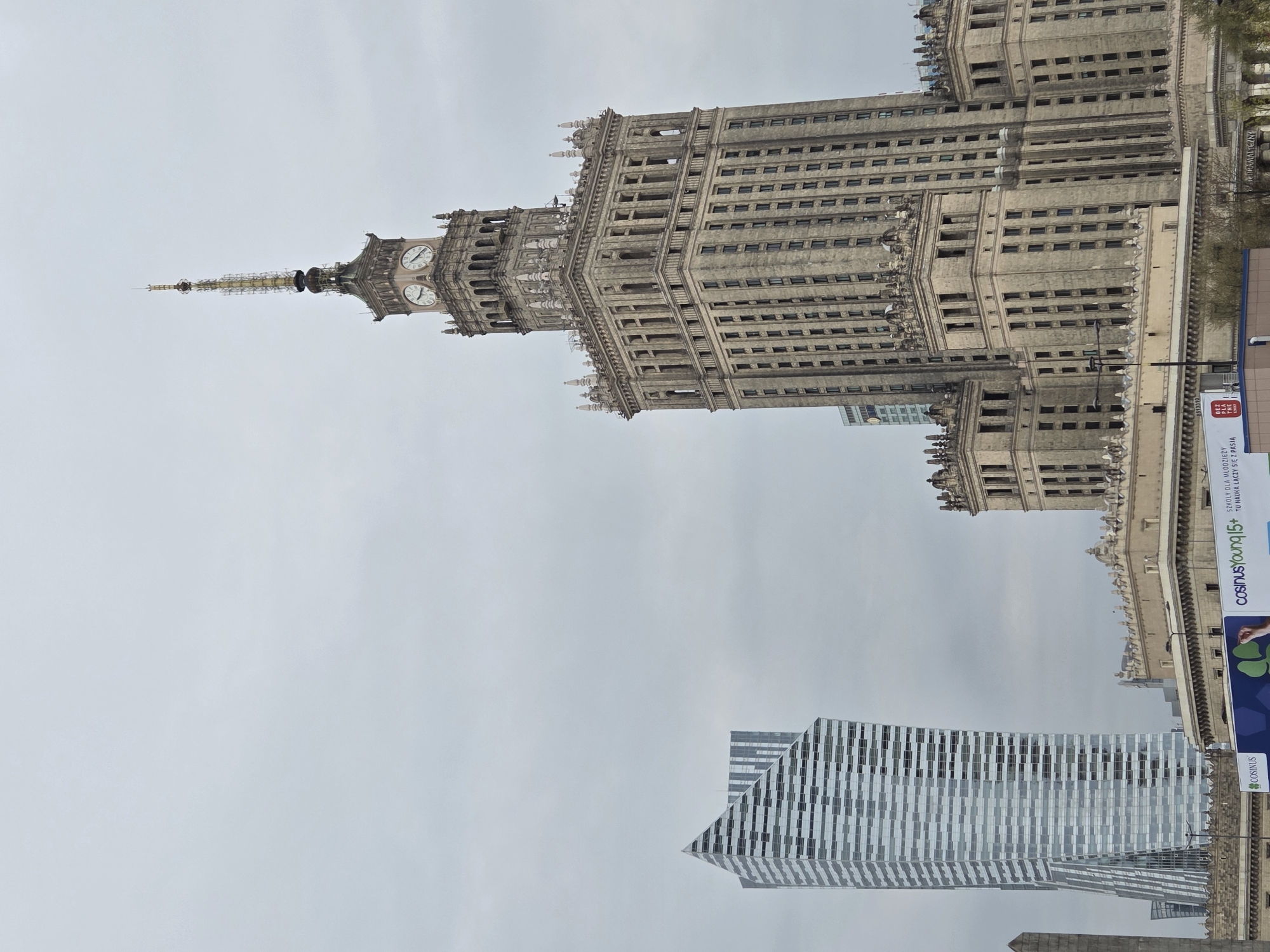 This image captures the Palace of Culture and Science in Warsaw, Poland, with a modern office building in the foreground. The sky is overcast, and the spire of the Palace of Culture appears to be under construction or renovation. A large banner is visible along the right side of the frame.