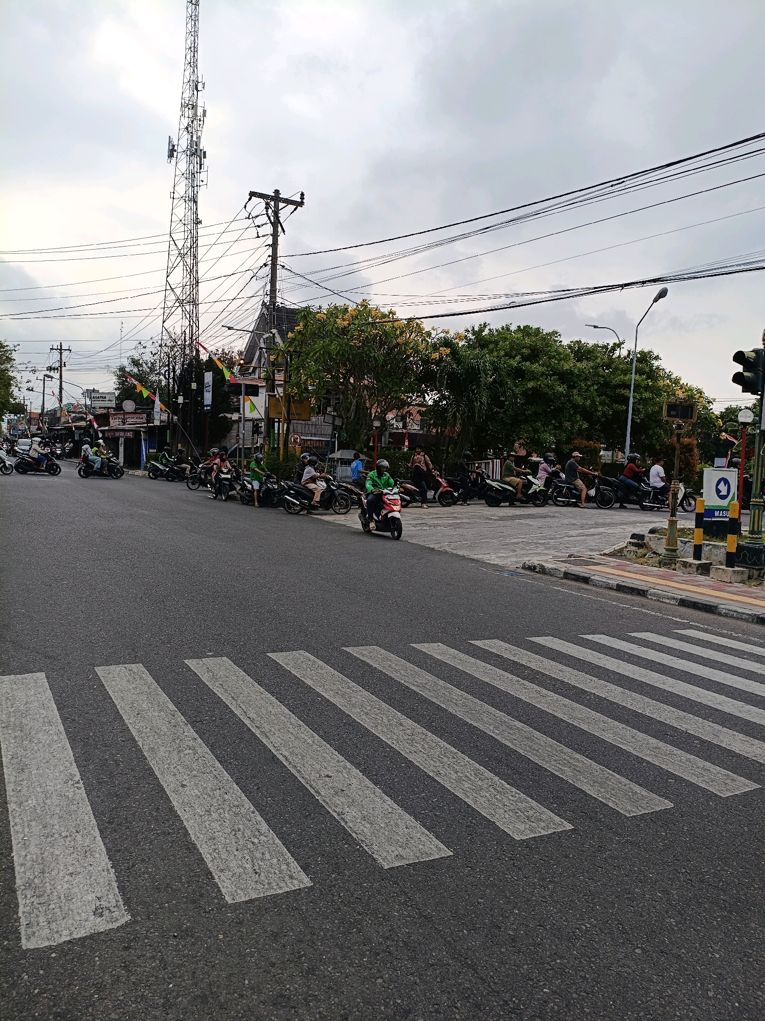 The image shows a street scene in the city of Yogyakarta, Indonesia, with a pedestrian crossing in the foreground. Several motorcycles are parked or waiting at an intersection. A tall telecommunications tower stands prominently in the background, with utility poles and power lines crisscrossing the sky. Trees and buildings line the street, and flags are visible on some of the buildings. The sky is overcast, suggesting a cloudy day.