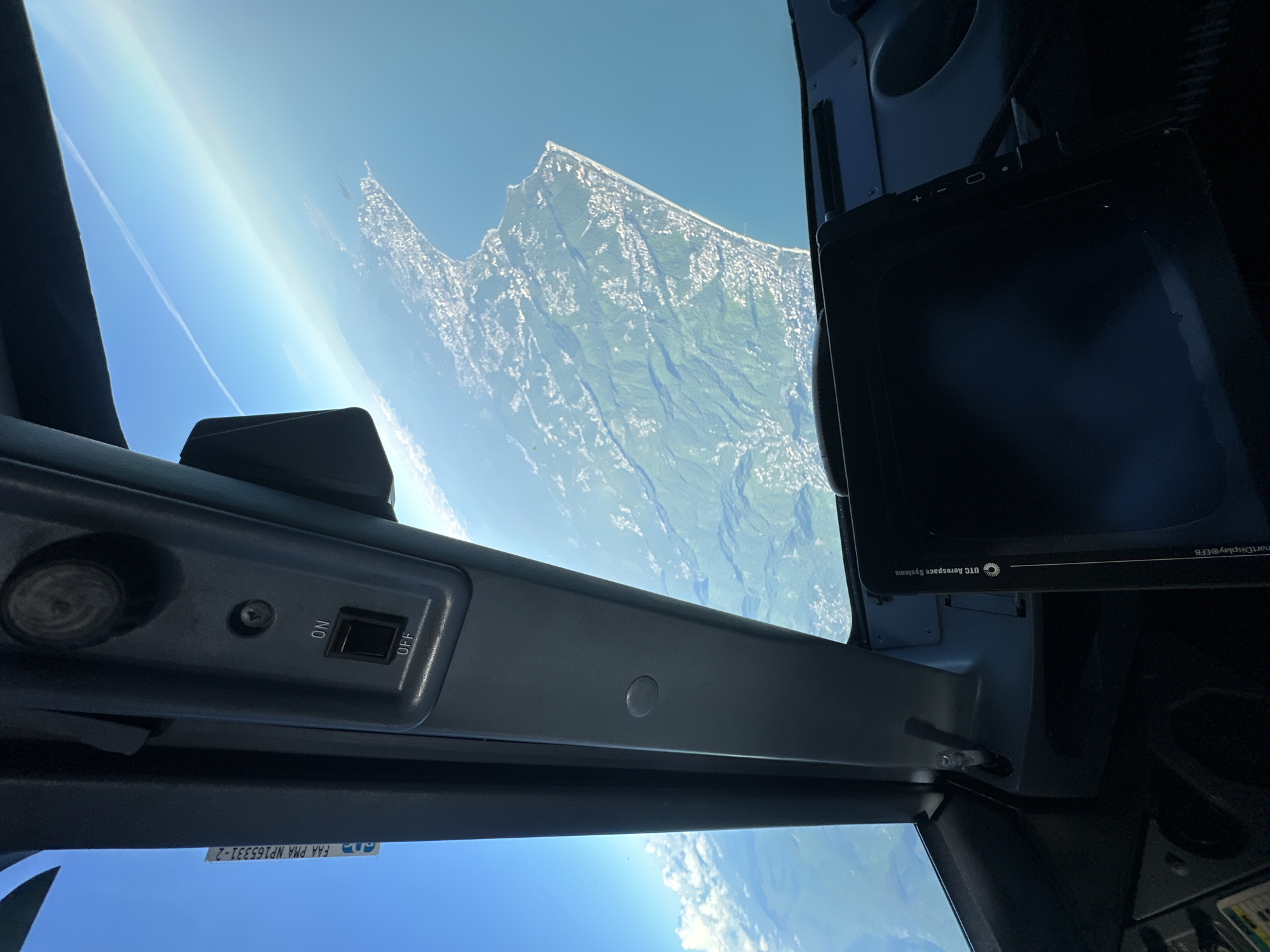 A view from inside an airplane cockpit shows a landscape outside the window. The landscape appears to be a combination of mountainous terrain with snow-covered peaks and green valleys, viewed from above. A contrail is visible in the sky. In the foreground, elements of the cockpit are visible, including a switch, a dark screen, and various buttons and controls. The city of Khimki, Russia, is not discernable in the image.