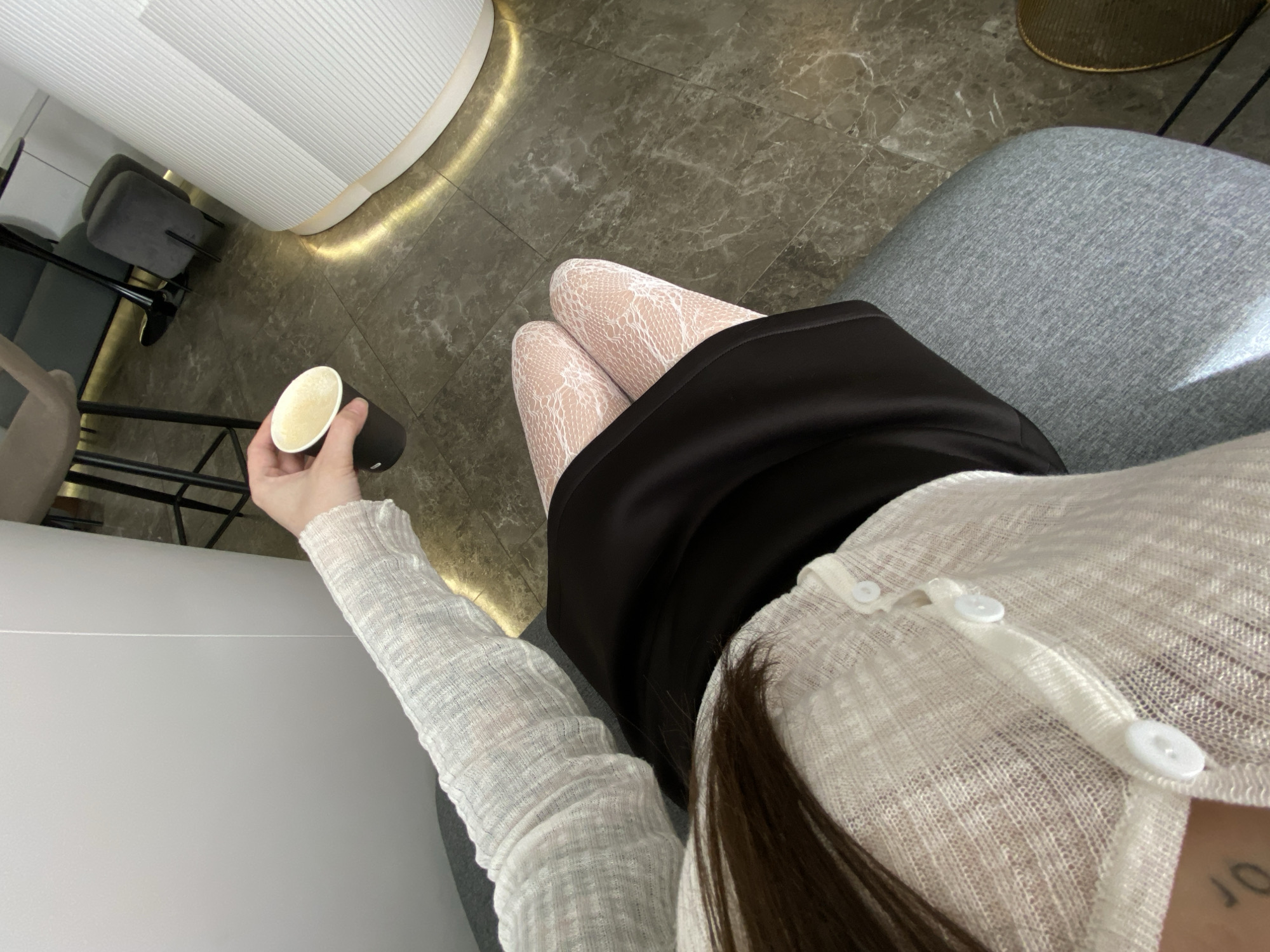 A high-angle shot captures a seated individual holding a beverage in a black cup, seemingly in an indoor setting. The person is wearing white lace stockings under a black skirt, paired with a long-sleeved white cardigan secured with white buttons. The individual is seated on a gray upholstered chair. The background includes a gray tiled floor, a ribbed white structure, and the legs of a metal chair. The setting's location is unspecified, and its relation to Votkinsk, Russia, is unknown.