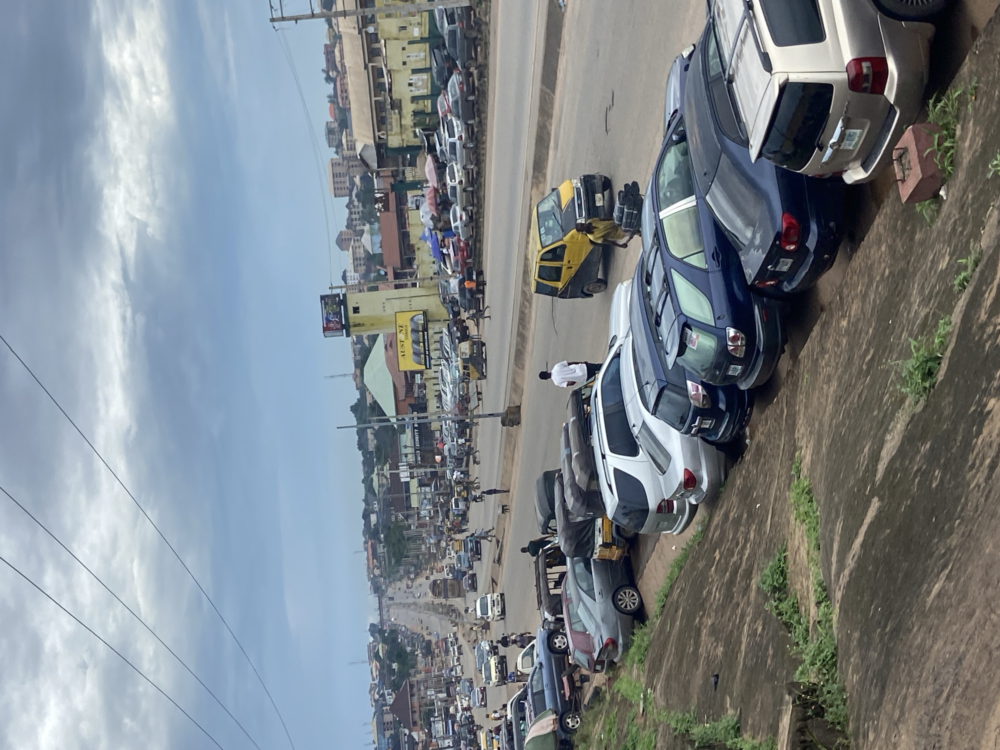 The image depicts a street scene in Ogbunike, Nigeria, characterized by a moderately busy road with several cars, mini-vans, and a yellow and black van. The road is lined with parked vehicles, predominantly on the right side, and features pedestrian activity. Buildings, including commercial establishments with signage such as "AUSTINE TIRES" and "SECURITY D," are visible along the roadside. The environment appears to be a commercial area with an urban landscape. The sky is overcast with clouds. The foreground shows a rocky incline with some vegetation.