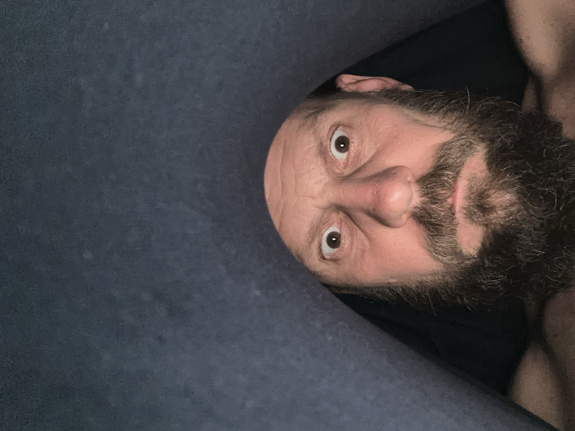 A man's face is closely framed within an opening in what appears to be a blue fabric or material. His eyes are wide open and looking directly at the viewer. He has a full, dark beard with some gray hairs mixed in. The background outside the opening is dark. The image provides insufficient context to identify a location in Fethiye, Turkey.