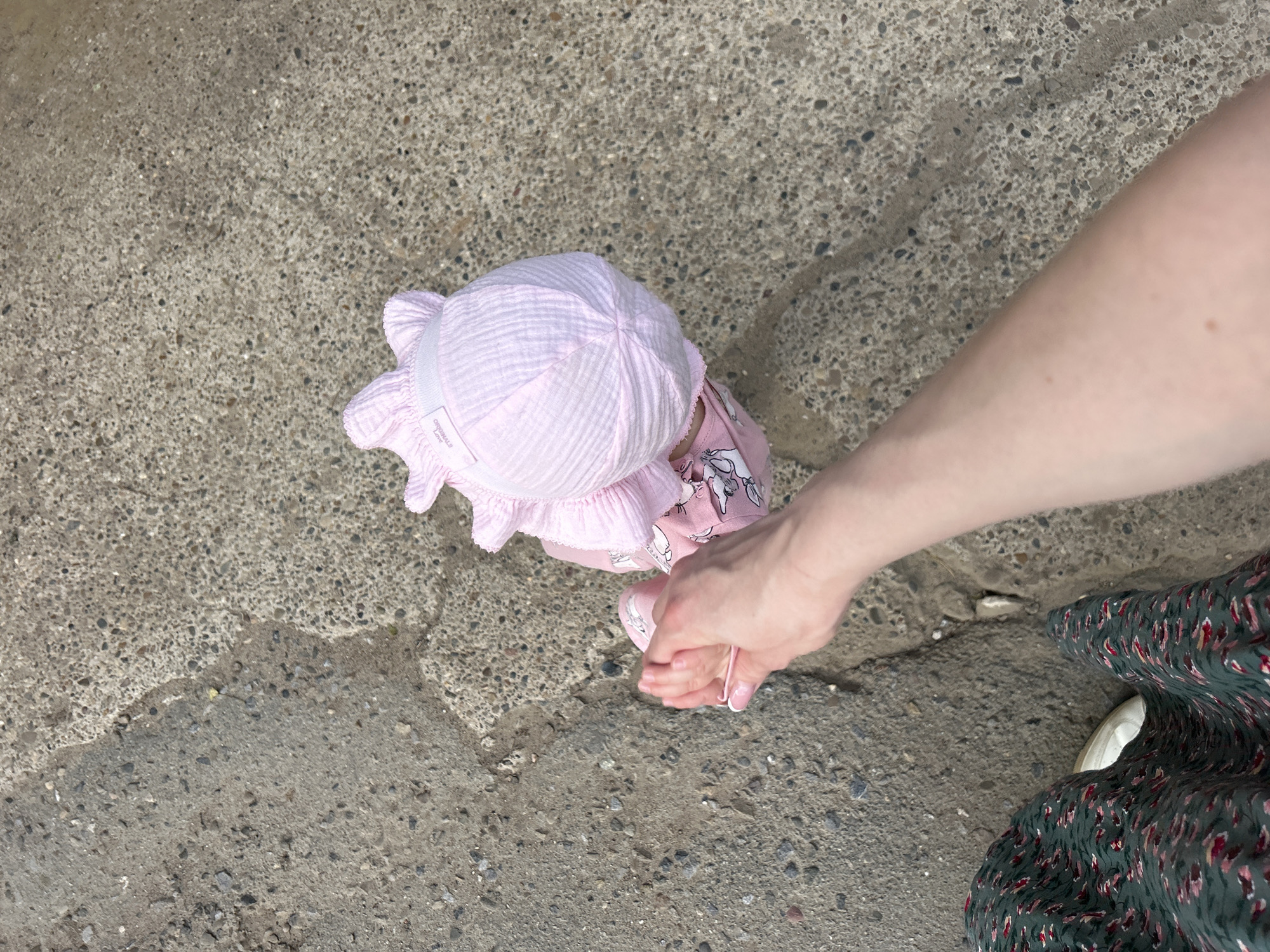 An overhead view shows a person's arm extending down to hold the hand of a baby wearing a pink sun hat and pink clothing. The baby is facing away from the camera and appears to be standing on a concrete surface that shows signs of wear and cracking, with some patches of dirt and debris. Part of a green floral patterned dress and a white shoe are visible in the lower left corner of the frame, indicating the presence of another person, likely the one whose arm is reaching down. The scene appears to be outdoors, on a paved area that could be a sidewalk or pathway in a city. The lighting suggests daytime, possibly overcast conditions.