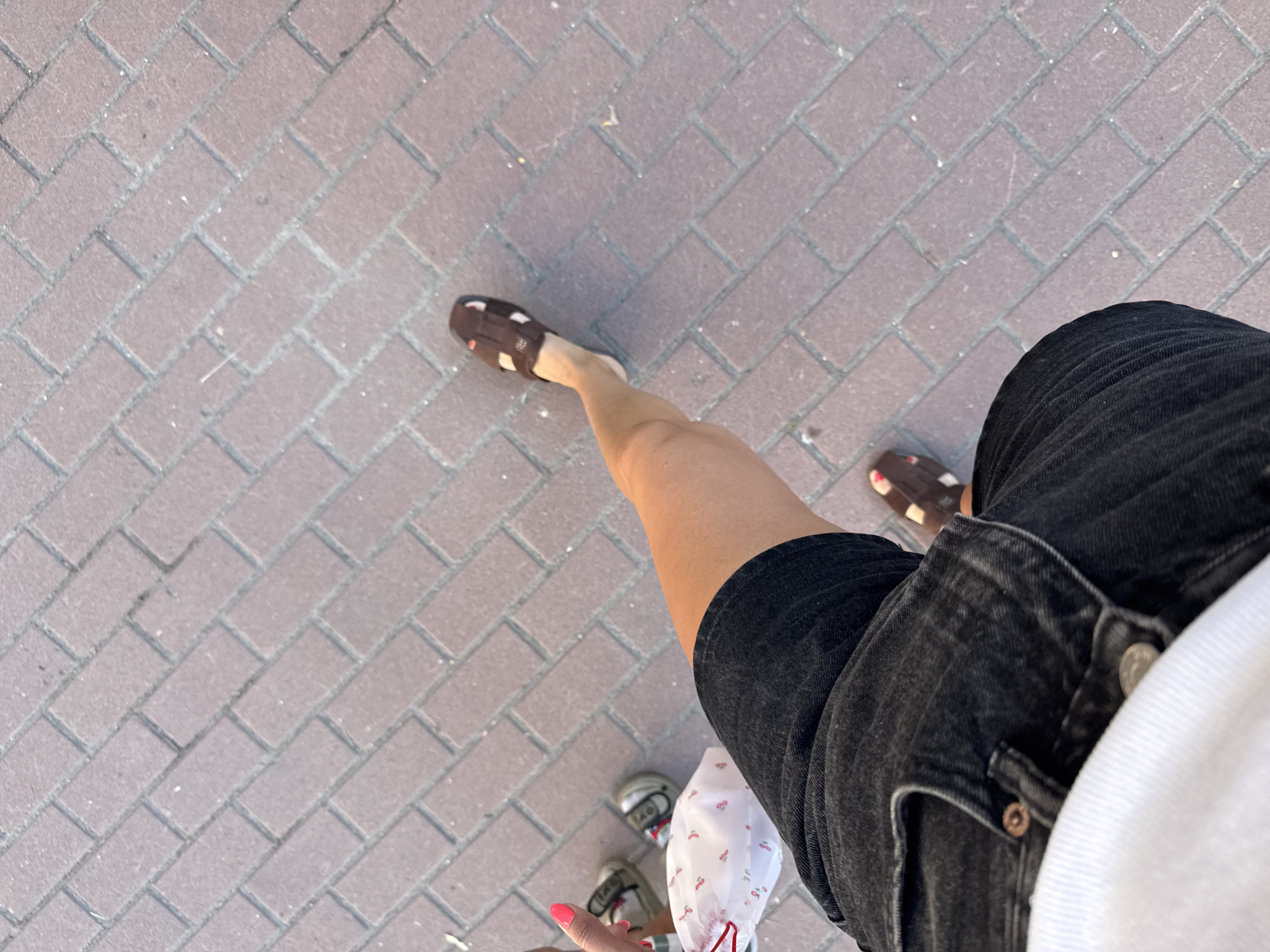The image captures a person's legs from a high-angle perspective, walking on a brick-paved sidewalk. The person is wearing black denim shorts, a white top, and brown sandals. In the background, two small children's feet are visible, also on the sidewalk, wearing small sneakers. The brick pavement is laid in a diagonal herringbone pattern. The scene appears to be a casual outdoor setting in a city, though no specific landmarks identifying Kyiv, Ukraine, are present. The lighting suggests it is daytime.