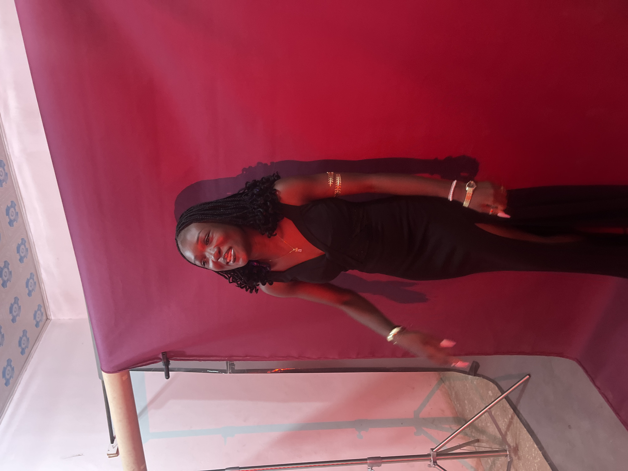In Gura Top, Nigeria, a young Black woman stands in front of a dark red backdrop, likely in a studio. She is wearing a sleeveless black dress with a thigh-high slit. Her hair is styled in braids, and she has on gold armbands on both upper arms, a necklace with a pendant, a watch, and rings on her fingers. Her mouth is slightly open, and she appears to be laughing or speaking. To the left of the woman, a photography stand with a light is visible, suggesting a professional or semi-professional photo shoot setup. The ceiling above has a decorative pattern.