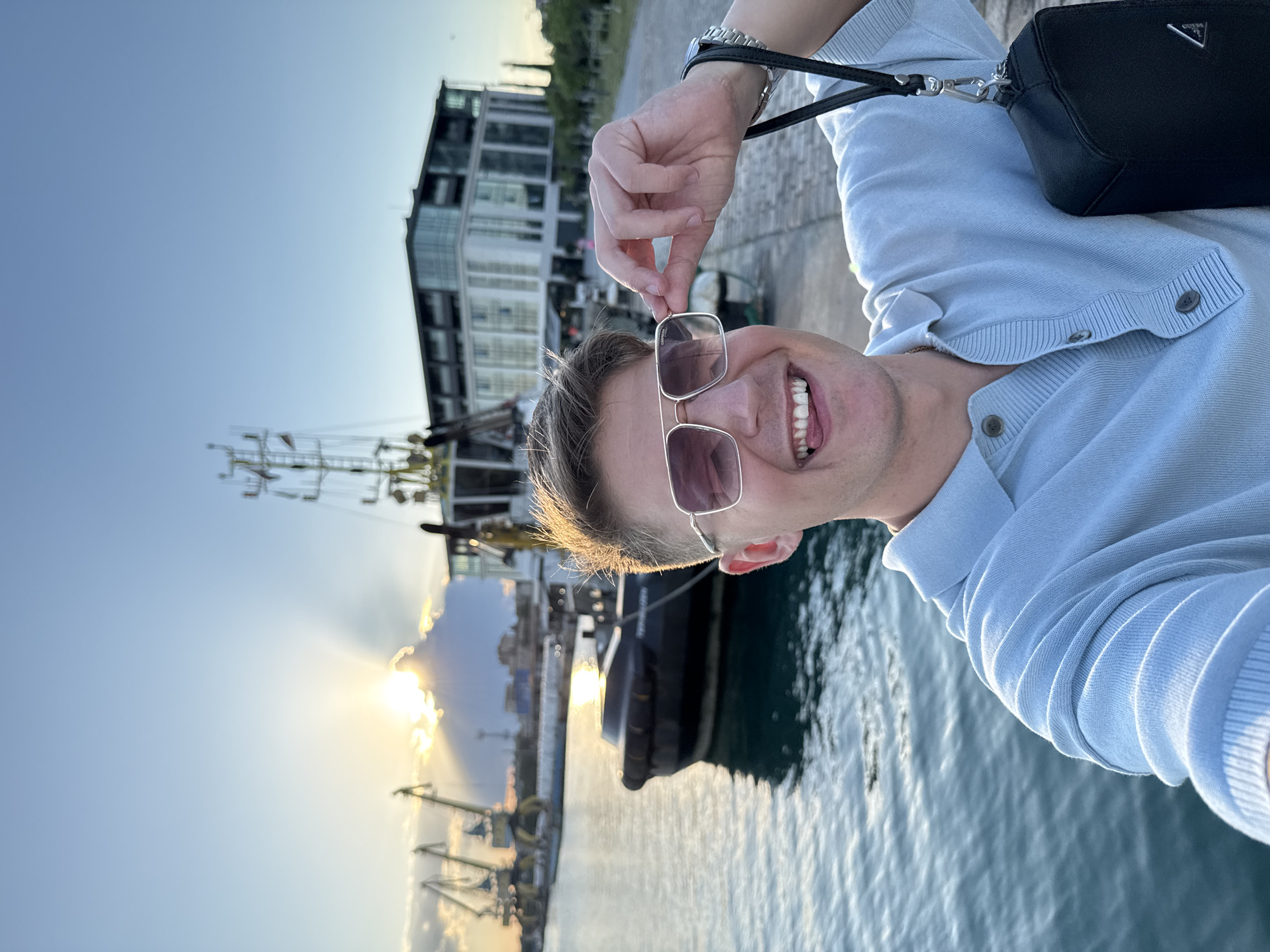 A man is taking a selfie at sunset by the sea in Burgas, Bulgaria. He is wearing sunglasses and a light blue polo shirt, and is holding a black bag. In the background, there is a tugboat, cranes, and a modern building. The sun is setting behind clouds, casting rays of light across the sky. The water is calm and reflects the sunlight.