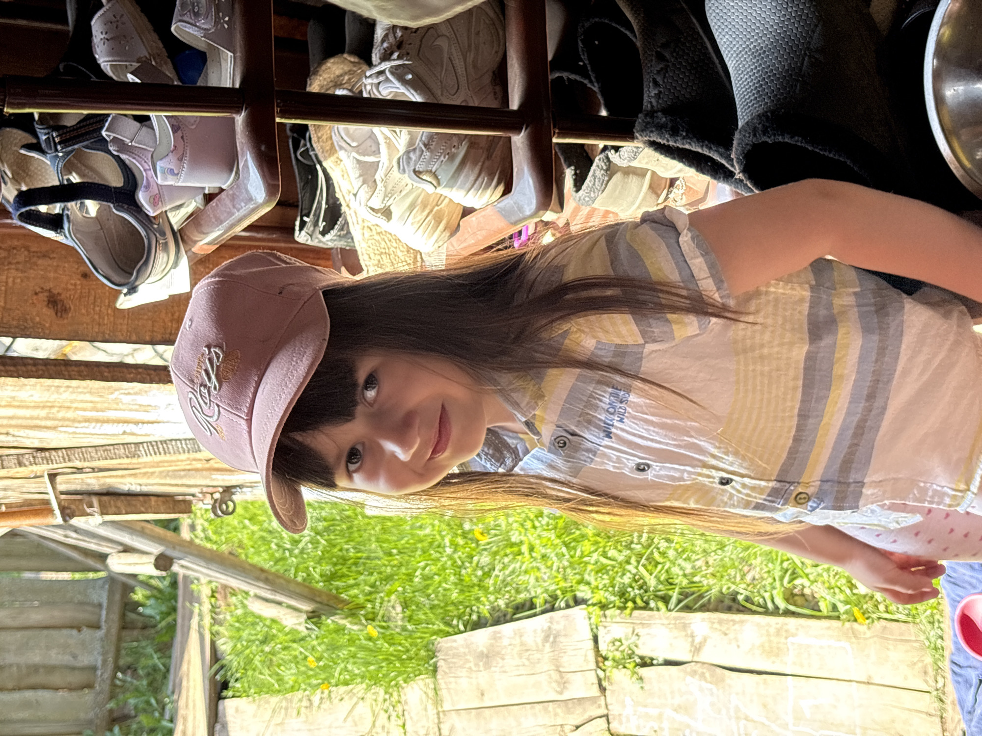 A young girl with long, dark hair and bangs is wearing a pink baseball cap and a striped shirt. She is looking at the camera with a slight smile. She appears to be outdoors, standing near a wooden structure with grass and foliage visible in the background. Above the girl, there is a shelf filled with various shoes. The location is not identifiable as Rosstani, Russia.