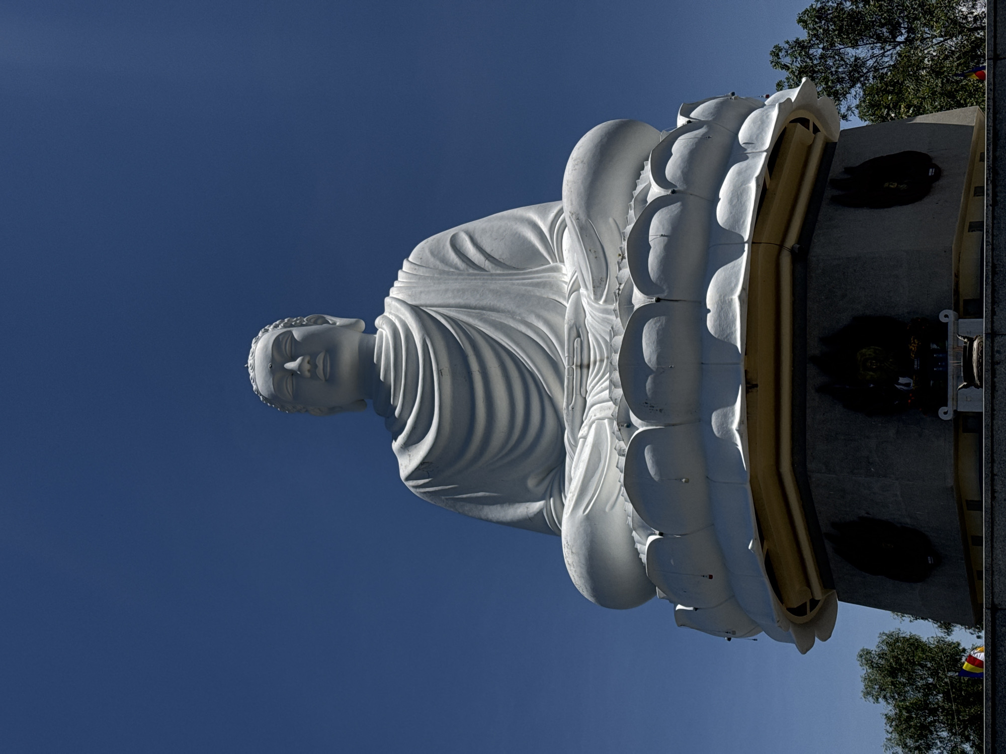A statue of a seated Buddha with a serene expression is oriented on its side, surrounded by a lotus flower pedestal against a clear blue sky. The statue is white, with gentle folds and curves defining the robe and body. The lotus flower features prominent, round petals. Behind the statue, a concrete structure with dark indentations is visible, with some foliage and a flag in the corner. The scene is the Long Son Pagoda in the city of Nha Trang Ward, Vietnam.