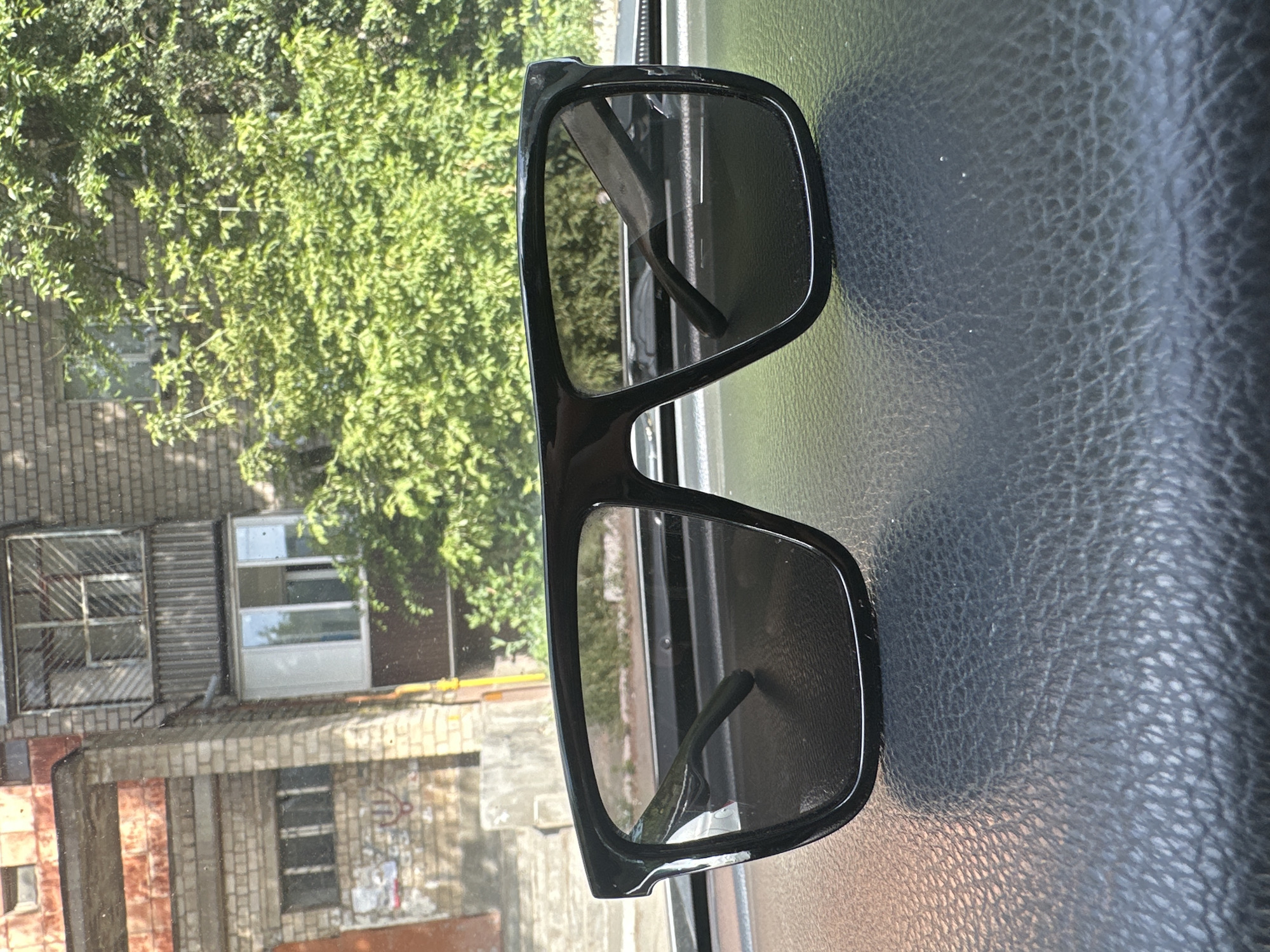 A pair of black, square-framed sunglasses with dark lenses rests on a dark, textured surface, possibly a car dashboard, with the lenses reflecting a bright outdoor scene. Behind the sunglasses, a building with brick and concrete elements is visible, along with lush green trees and foliage. The building appears to be a multi-story structure with balconies and windows, suggesting an urban or suburban environment. The sunlight indicates it is daytime. The overall setting, with the building and trees, is consistent with the general appearance of residential areas in Orsk, Russia.