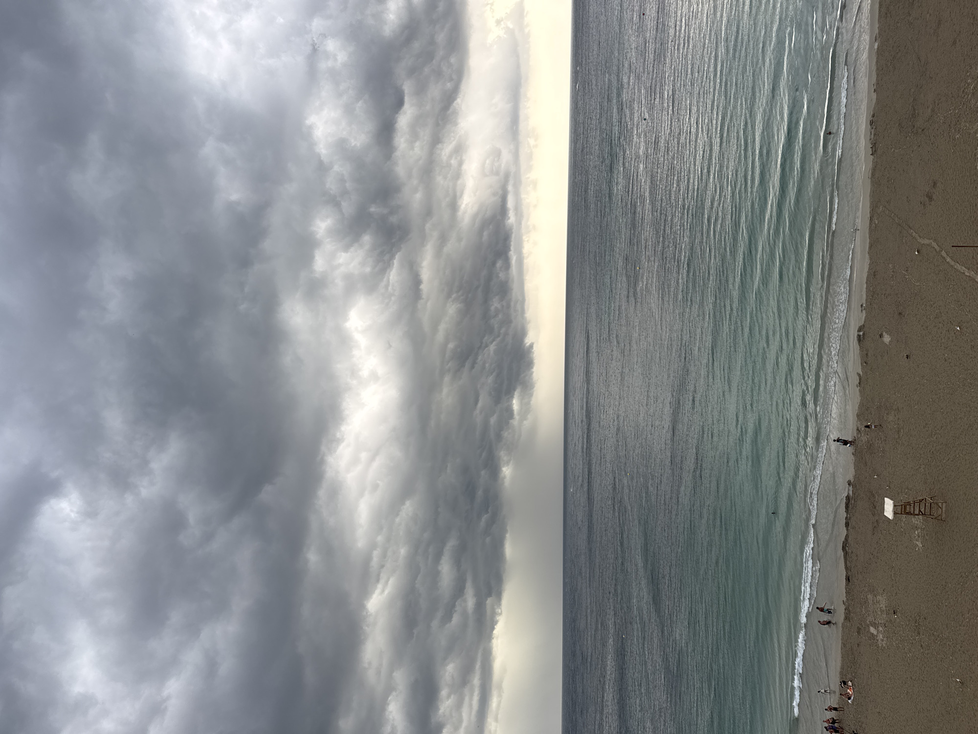 The image depicts a cloudy day at a beach in Calp, Spain. The sky is dominated by heavy, dark gray clouds, with a lighter, overcast sky visible near the horizon. Below the sky, the ocean stretches out, with the water transitioning from a dark, deep blue in the distance to a lighter, turquoise hue closer to the shore. Gentle waves lap at a sandy beach.
On the beach, several people are present. In the foreground, the sand is mostly empty, with a raised lifeguard chair standing prominently. Further down the beach, closer to the water, small groups of people are scattered. Some are walking along the shore, while others appear to be relaxing or playing in the shallow water. One person is visible swimming in the ocean.
The overall atmosphere is subdued, likely due to the overcast weather, but the presence of people suggests that activities on the beach continue despite the clouds. The horizon is clearly defined, separating the sea from the sky.