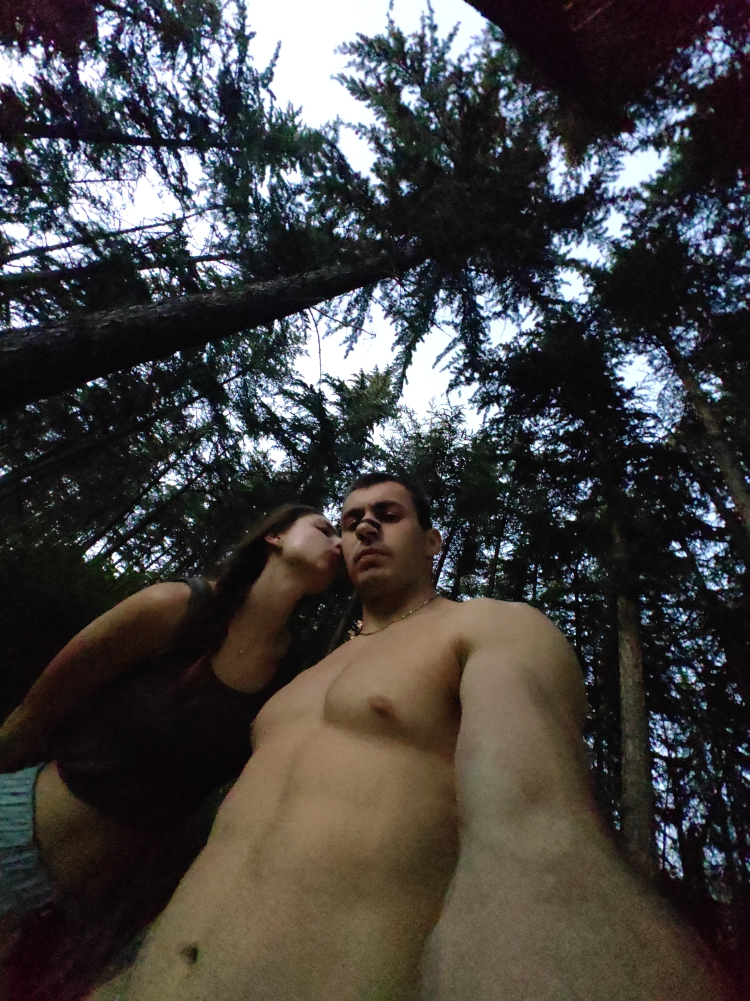 An outdoor, low-angle, wide shot captures two individuals, a man and a woman, in a forest setting. The man, shirtless and in the foreground, is facing the camera with his head slightly tilted. He has short dark hair and a gold chain around his neck. A dark, vertical mark is visible on his forehead. The woman is positioned to his left, leaning in to kiss him. She has long brown hair and is wearing a dark top. The background is dominated by the dense canopy of tall trees, with glimpses of the sky visible through the branches. The lighting suggests it is either dusk or dawn. The image does not provide any specific visual cues to identify a precise location within the city of Varna, Bulgaria, other than it being a forested area.