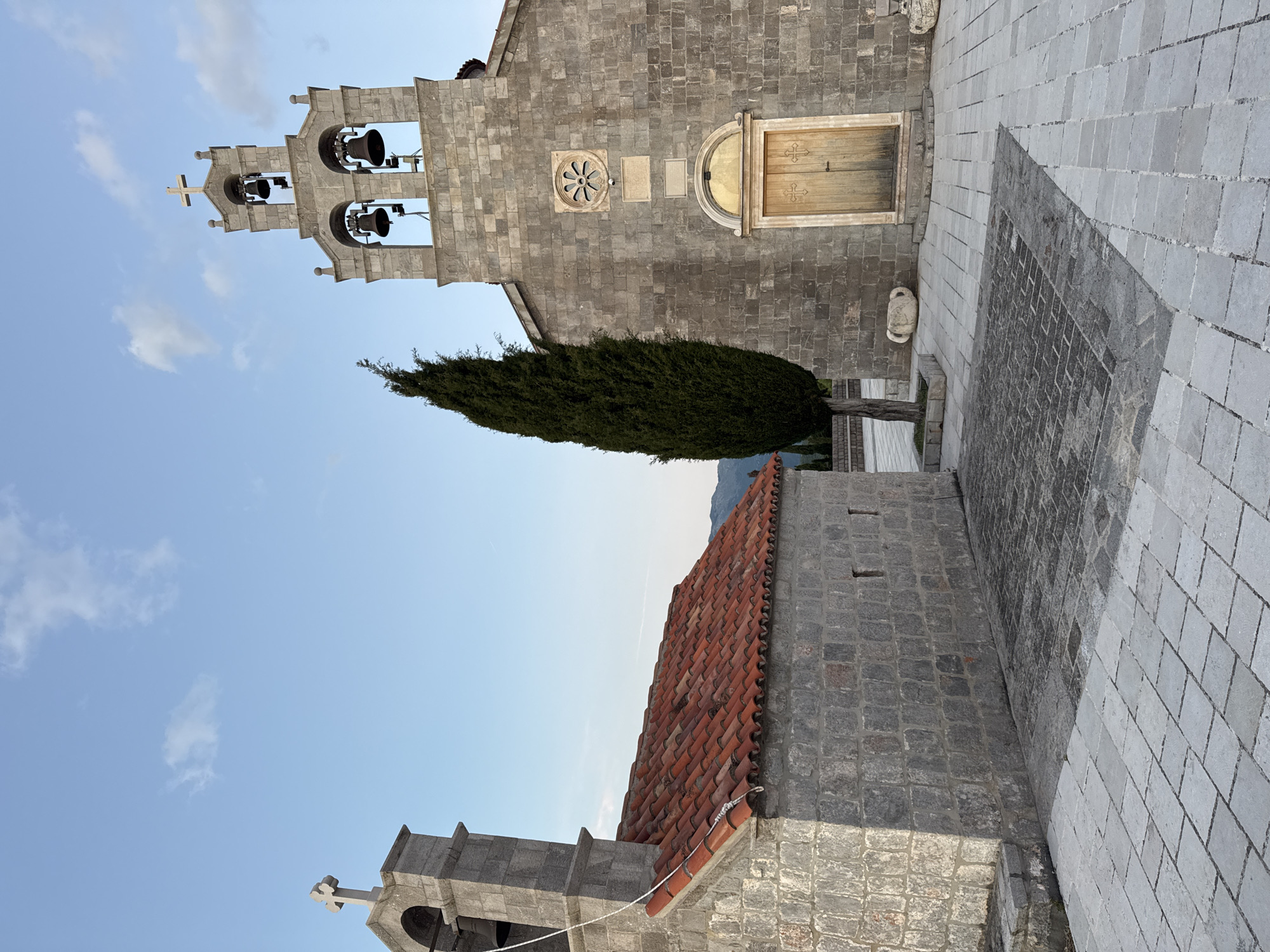 A rotated, medium shot depicts two stone buildings, a tall, dark green, cypress-like tree, and a stone-tiled plaza set against a blue sky with scattered clouds.
The larger, older building features a bell tower with multiple bells and a cross at the top. The building itself is constructed of gray stone blocks and includes a round, carved decoration and a wooden door.
A smaller, adjacent building has a red-tiled roof and a smaller stone bell tower, also with a cross.
The stone-tiled plaza extends in front of the buildings.
The scene is located in Kovačko Polje, Montenegro, as specified.