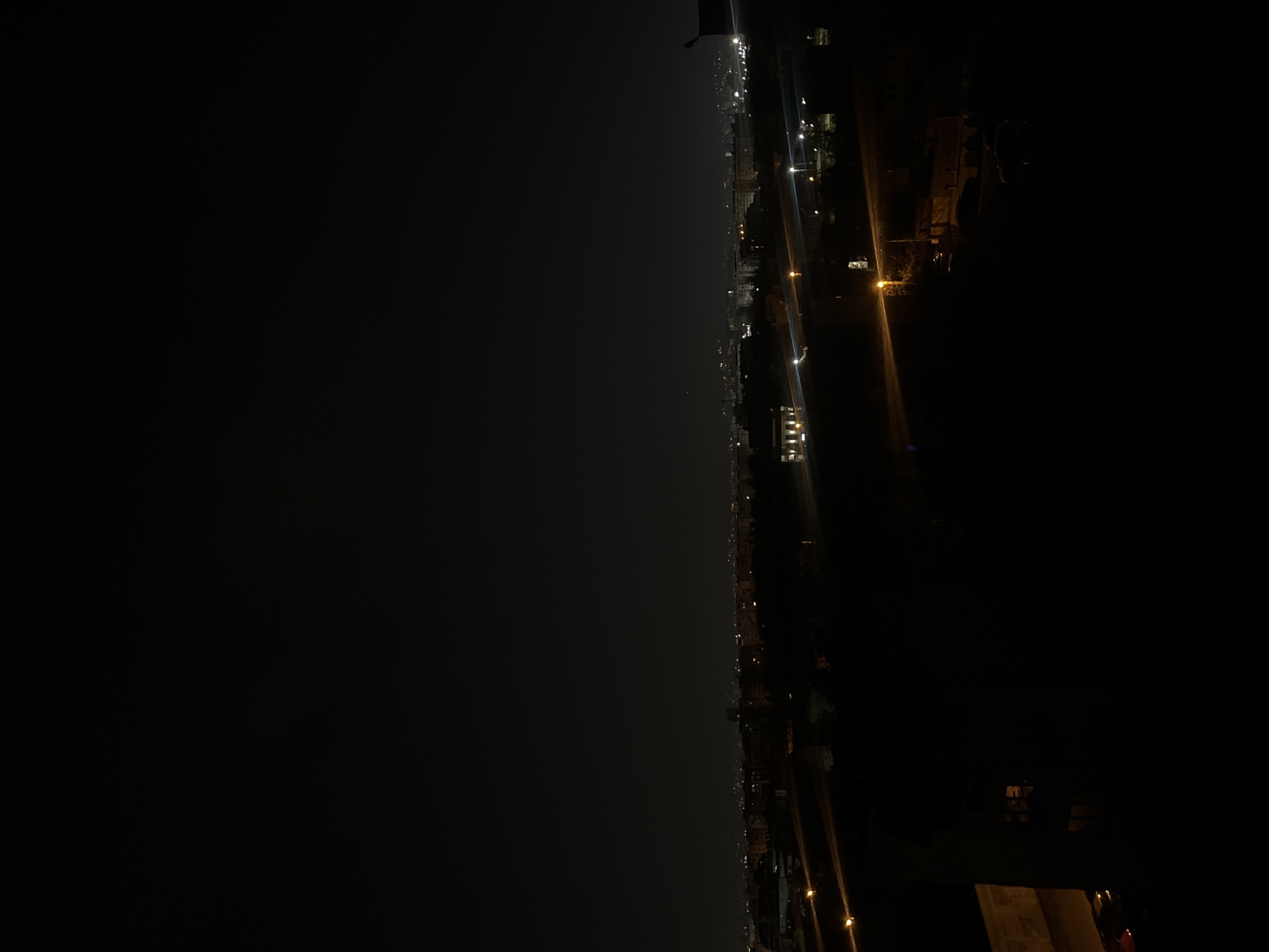 The image shows a night cityscape, likely taken from a high vantage point, potentially an upper floor of a building. The lighting is poor, with most of the details obscured by the darkness.
On the right, there are blurred lights, possibly from streetlights or buildings, indicating a road or urban area. In the central area, more dispersed lights from buildings can be seen.
The left side of the image has significantly fewer details, suggesting a vast open area, possibly a lake. Due to the limited visibility and information in the image, it is impossible to pinpoint the exact location within Sofia, Bulgaria or to identify specific subjects or actions.