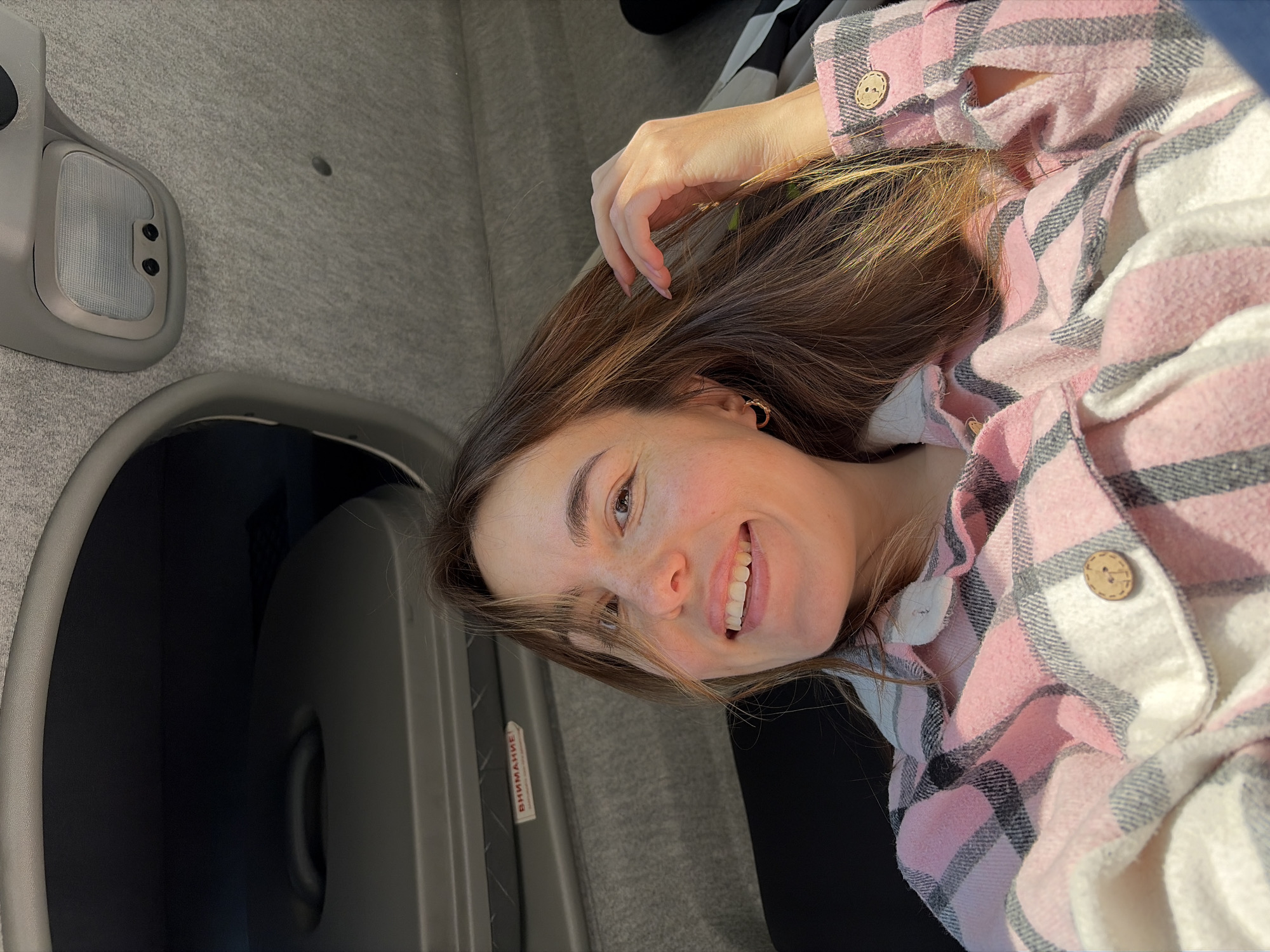 A young woman smiles at the camera. She is wearing a pink and white plaid shirt and has long brown hair. She is positioned inside a vehicle, near the roof of the interior of the vehicle with a ceiling light. There is a grey box nearby that contains a label with Cyrillic text. The Russian city is Izhevsk.