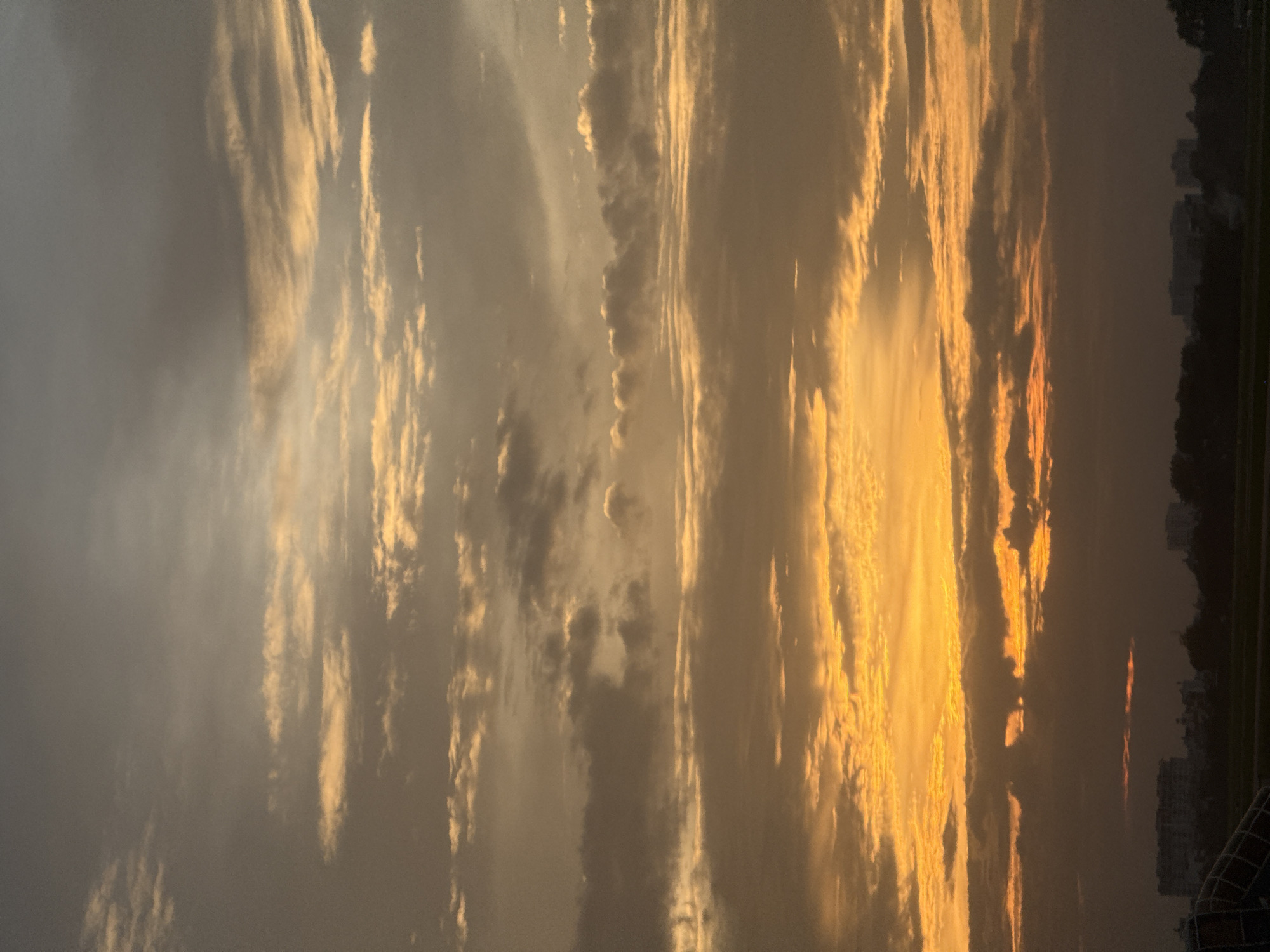 The image depicts a sunset over a cityscape in Ho Chi Minh City, Vietnam. The sky is filled with clouds, ranging in color from dark gray to vibrant orange, indicating the sun is low on the horizon. Buildings and trees are visible in silhouette against the bright sky at the bottom of the image. The overall scene is one of natural beauty as the day transitions into night.