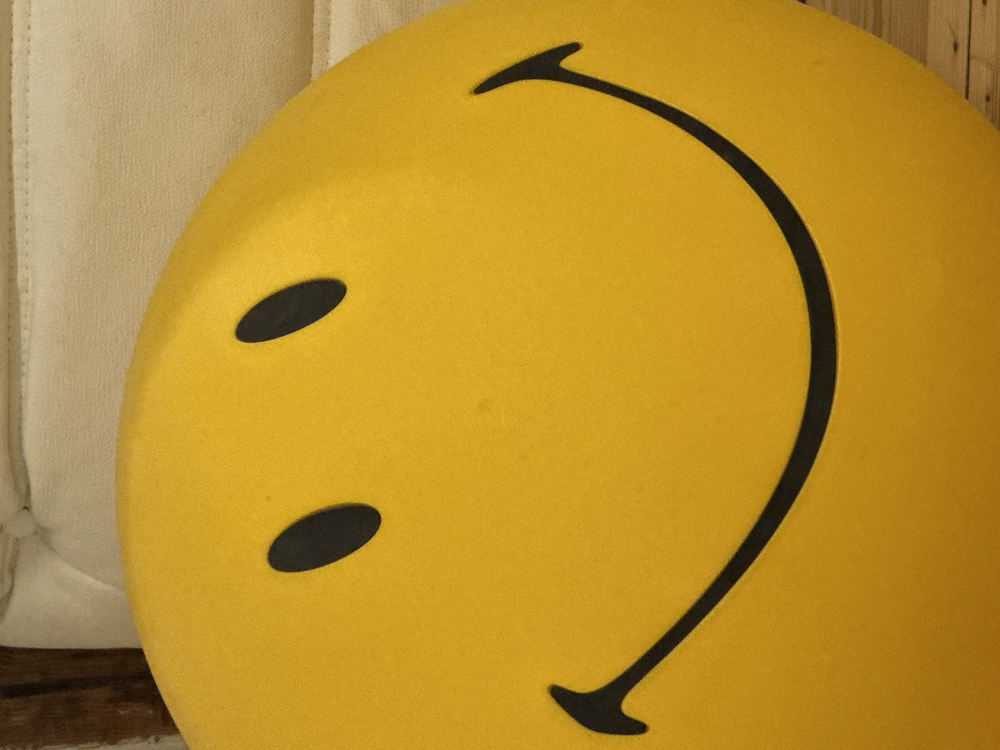 A close-up photograph captures a large, bright yellow, circular object that resembles a classic smiley face. The object dominates the frame, positioned towards the right. It features two prominent black, oval-shaped eyes and a wide, upward-curving black smile, all rendered in a bold, graphic style. The yellow surface has a fine, slightly textured appearance.
To the left of the smiley face, a portion of a light-colored, possibly cream or beige, textured fabric object is visible. This fabric appears to be upholstered, with subtle vertical stitching, suggesting it could be part of a cushion, chair, or sofa. Hints of a wooden surface, possibly planks, are also visible along the far right and bottom edges of the image, indicating an indoor setting.
There is no activity or event taking place; the image is a static shot of an inanimate object. No people are visible. The object itself conveys an emotion of happiness or cheerfulness due to its design. There is no visible text in the image. The setting is generally indoors, located in London, United Kingdom.