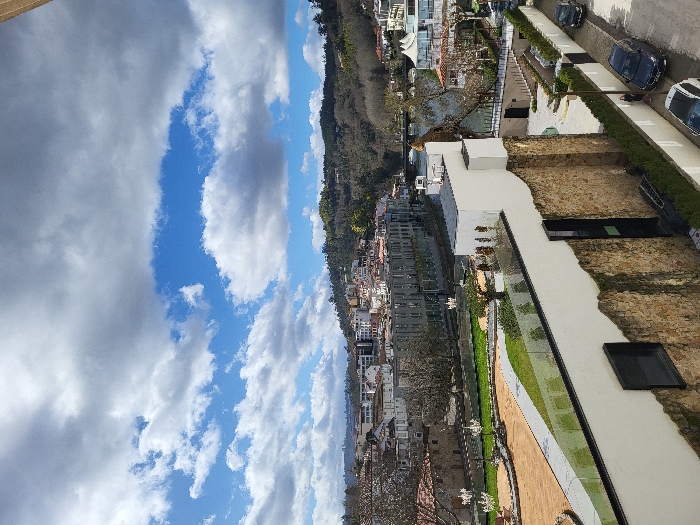 An elevated, wide-angle photograph captures a cityscape in São Pedro do Sul, Portugal, under a partly cloudy sky featuring prominent white and grey cumulus clouds against a blue backdrop.
In the immediate foreground, the upper section of a multi-story building facade is visible. It features a rough, irregular stone-block base supporting a smooth, white rendered wall. A clear glass railing tops this white wall, extending towards the left. A dark, rectangular window is set into the white wall, and a tall, narrow black-framed door or window is positioned to the right within the stone section. A silver ladder leans against the stone wall near this dark opening.
To the right of the foreground building, a paved street runs parallel, featuring a low green hedge. Three vehicles are parked along this street: a dark SUV closest to the camera, a dark sedan, and another dark vehicle further in the distance. A single individual is standing on the sidewalk next to the dark sedan.
Beyond the foreground structure, a landscaped park area unfolds. This area features sections of green grass, winding light-colored paved paths, and reddish-brown ground. Several multi-branched white outdoor chandeliers are suspended over this park. A dark body of water, identified as a river, flows from the right across the midground, with a stone-arched bridge spanning it further to the left.
On the opposite bank of the river, a dense urban area is visible. A large, grey, multi-story building with numerous windows dominates this section, flanked by other structures with white facades and red-tiled roofs. The background consists of verdant hills, dense with trees and bushes, dotted with additional residential buildings, rising under the open sky.