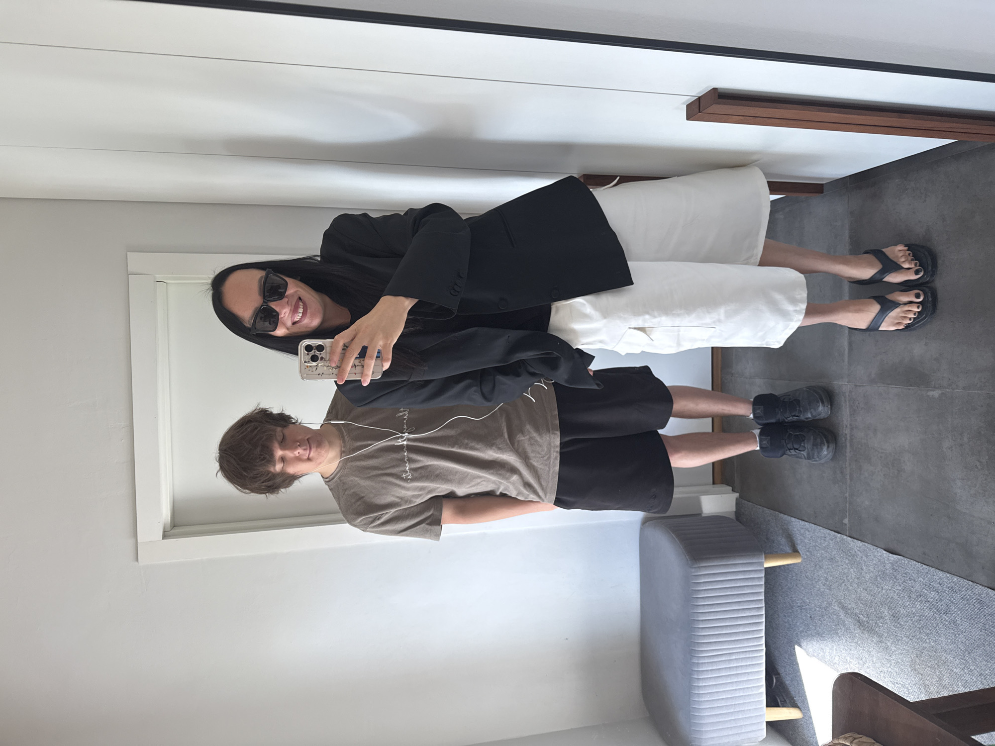 In what appears to be a residential setting, possibly an apartment hallway or dressing room in Chaikovsky, Russia, two individuals are captured in a mirror selfie. The person on the left, a young male with disheveled brown hair, wears a taupe t-shirt with a subtle graphic and black athletic shorts. White earbuds are visible in his ears. He stands next to a grey upholstered bench with wooden legs.
The person on the right, dressed in sunglasses and a black blazer over a black top, holds a smartphone and poses for the photo. They are wearing white, wide-leg cargo shorts and black thong sandals with dark nail polish. The background shows a white wall and a doorway. The floor is tiled in a dark grey. The overall lighting is bright, suggesting it is daytime.
