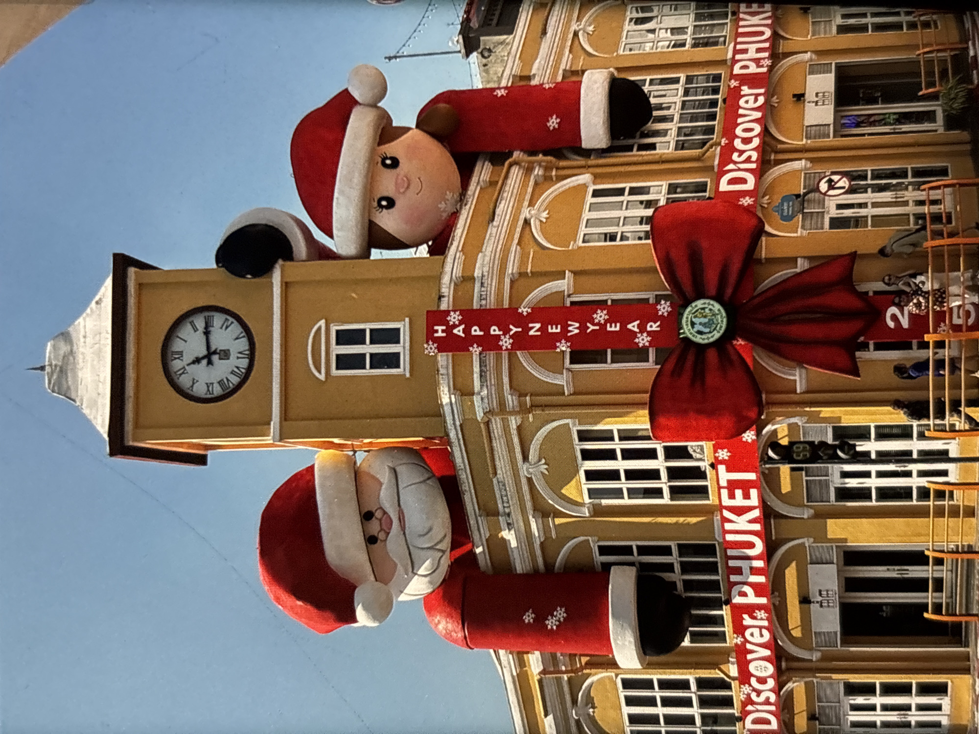 An outdoor, low-angle, full shot captures a brightly lit, yellow building adorned with large, festive Christmas decorations in Chalong, Thailand. Two oversized, inflatable figures, one resembling Santa Claus and the other a female character, both wearing Santa hats, peek over the building's upper facade. The Santa figure is on the left, and the female figure is on the right. The building features a prominent clock tower with Roman numerals on its face, positioned centrally above the figures. A red banner with white lettering that reads "HAPPY NEW YEAR" hangs vertically between the two inflatable figures. Below them, a large red bow is affixed to the building. Another red banner runs horizontally across the lower portion of the building's second story, displaying the white text "Discover PHUKET". The building has multiple windows and arched doorways. Several individuals are visible on the sidewalk in front of the building, including two women and a man. The sky is clear and blue.