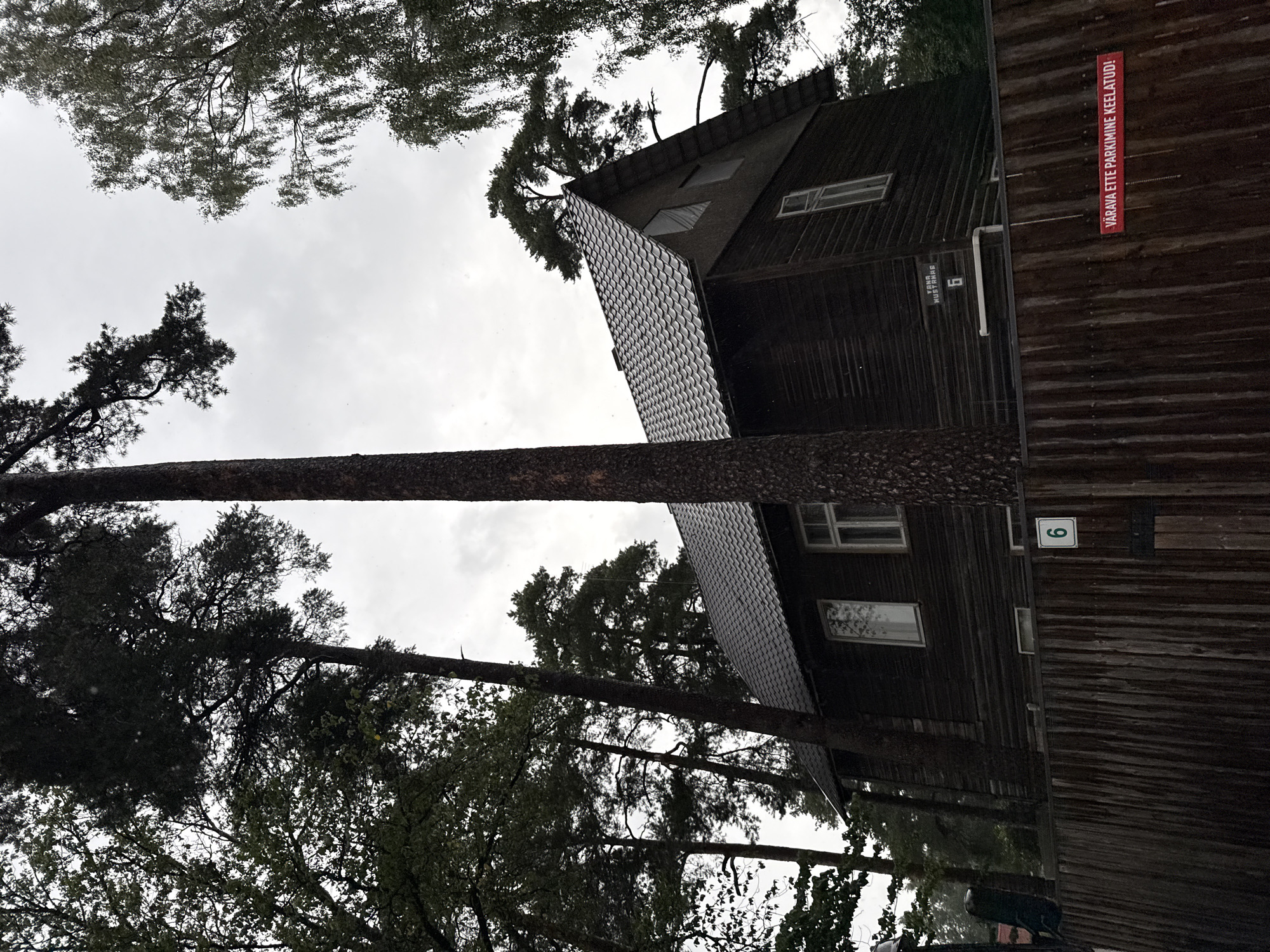 The image depicts a dark wooden house with a grey, shingled roof, partially obscured by tall trees. A wooden fence runs in front of the property, with a sign on the fence reading "VÄRAVA ETTE PARKIMINE KEELATUD!" which translates to "PARKING PROHIBITED IN FRONT OF THE GATE!". Another sign on the house itself displays the number "6" and text that appears to be "TAMA MUSTRAHG". A green sign with the number "6" is also visible on the fence. The sky is overcast, suggesting a cloudy or rainy day. The overall scene is residential, with dense foliage surrounding the building. The location is identified as Tallinn, Estonia.