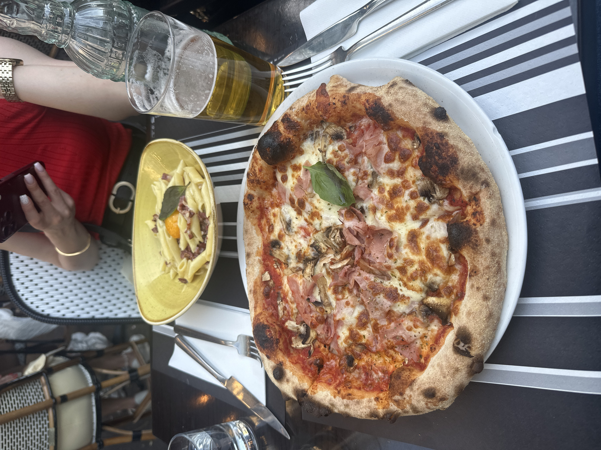A pizza and pasta dish are served on a black and white striped table. The pizza is round with a golden crust and toppings of ham, mushrooms, and cheese. A glass of beer sits next to the pizza. The pasta dish is yellow and contains penne pasta with a fried egg and bacon. In the background, a person wearing a red top and a gold bracelet holds a phone. A bottle of water is also visible on the table. The scene appears to be an outdoor dining setting in Paris, France, with bistro chairs in the background.
