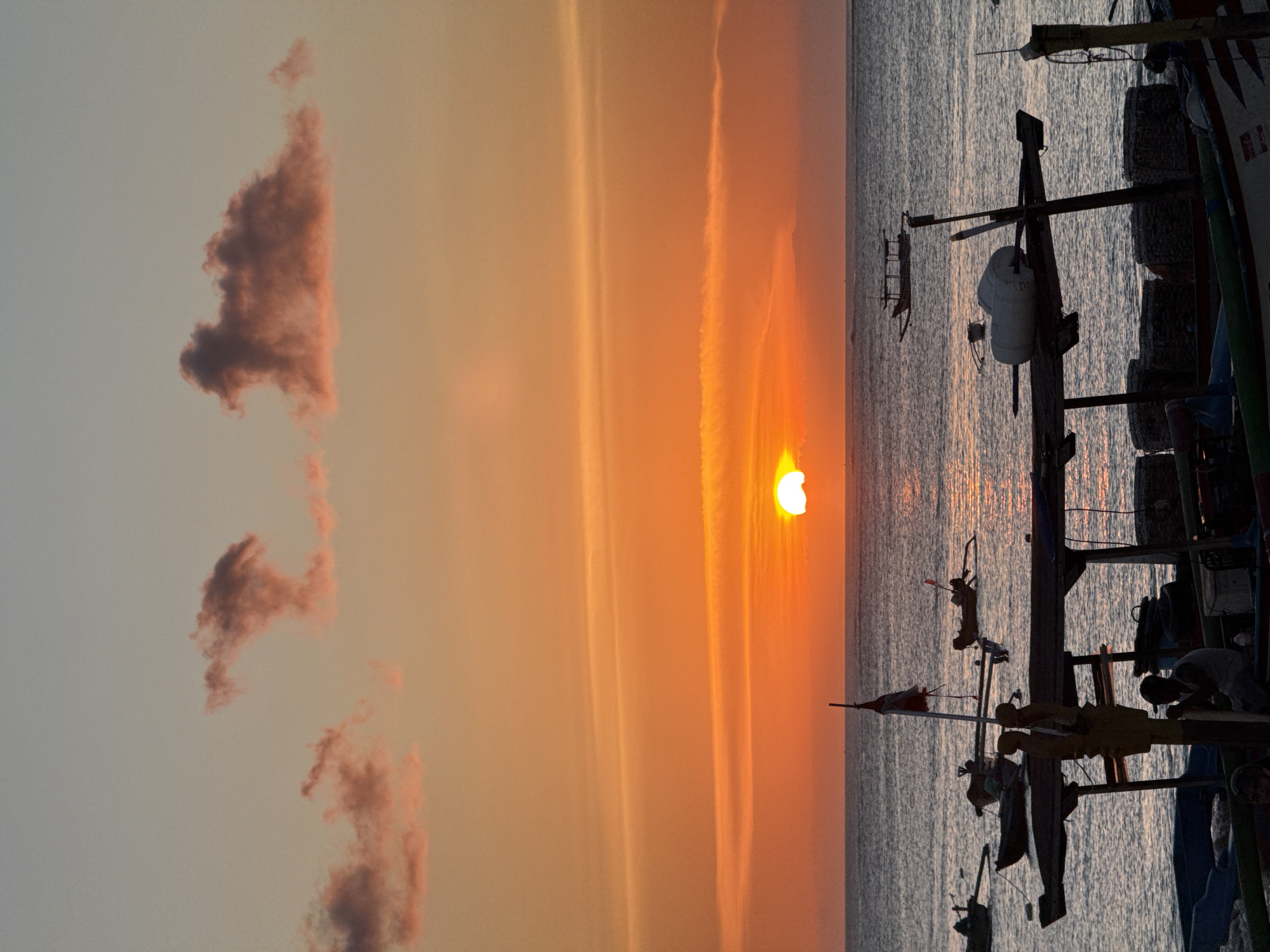 The image captures a sunset scene in Kuta, Indonesia. The sun is partially obscured by atmospheric haze and clouds, creating a warm, orange glow across the sky and reflecting on the ocean surface. Silhouetted against the sunset are traditional fishing boats, with their rigging and equipment visible. The boats appear to be docked or anchored close to the shore. There are some clouds in the sky.