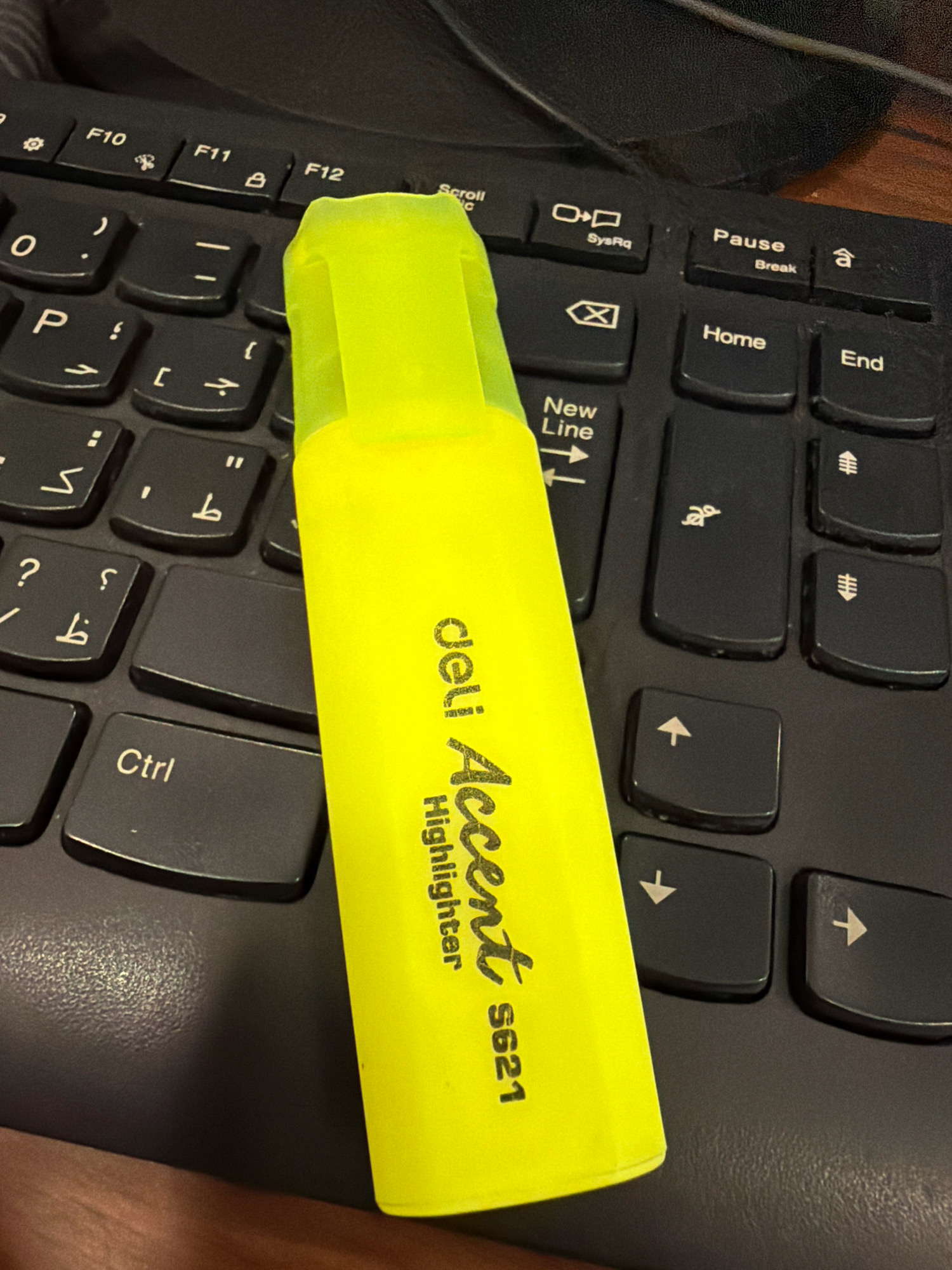 A bright yellow highlighter lies diagonally across a black computer keyboard. The highlighter is oriented from the bottom left to the top right of the image and has the text "deli Accent Highlighter S621" printed vertically on its side. The keyboard keys are visible beneath the highlighter, including "Ctrl", "New Line", "Pause", "Home", and "End". The image appears to be taken in a well-lit indoor environment, possibly an office or home workspace in Riffa, Bahrain.