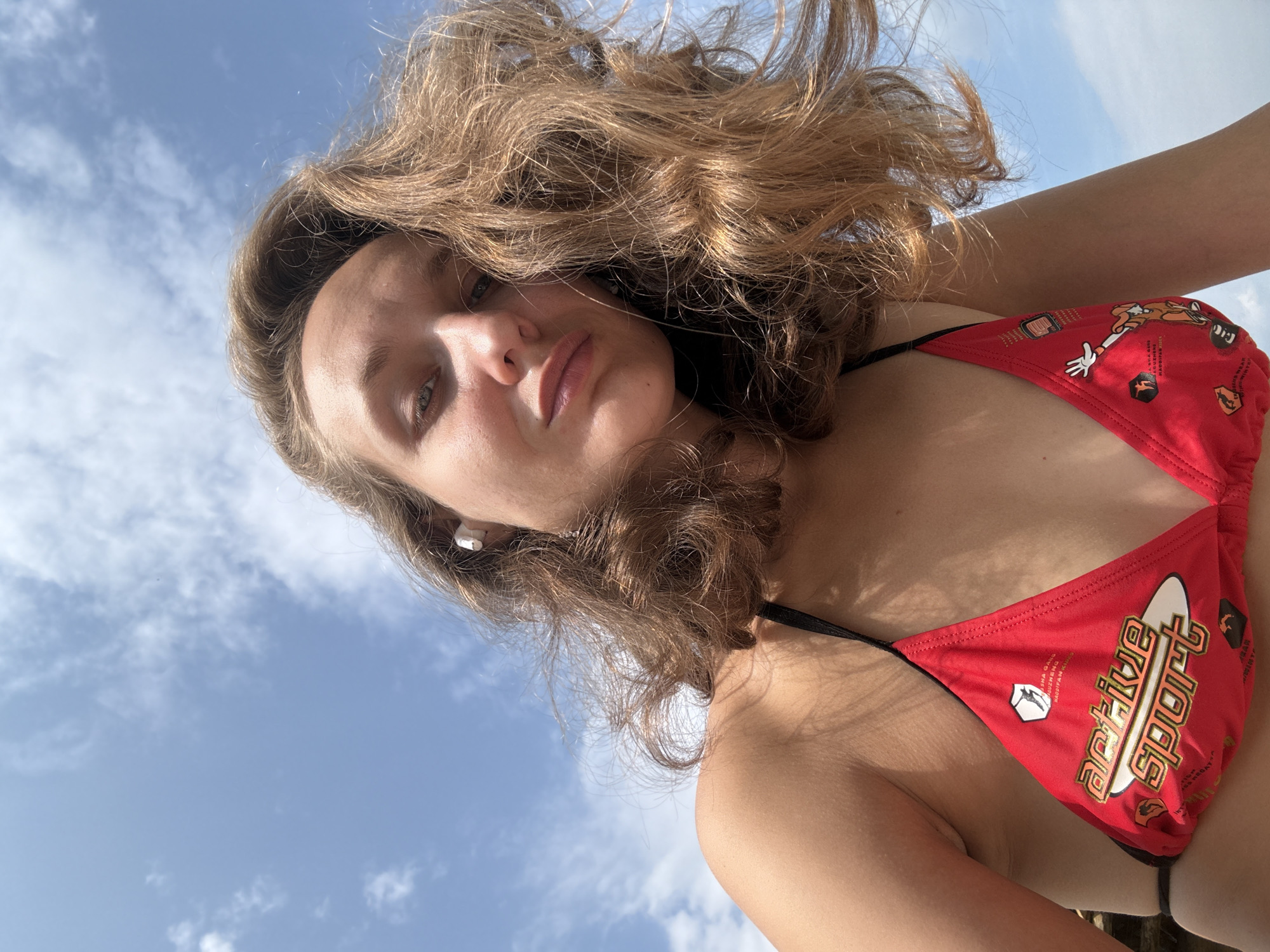 A low-angle, medium shot shows a young woman wearing a red bikini top with black trim. The bikini has a graphic print including the words "active sport" in white and gold. She has shoulder-length, wavy, light brown hair, and is wearing white wireless earbuds. She is looking at the camera with a neutral expression. The background is a bright blue sky with scattered white clouds. The image appears to have been taken outdoors on a sunny day. There is no visible indication of the location being Podshivalovo, Russia.